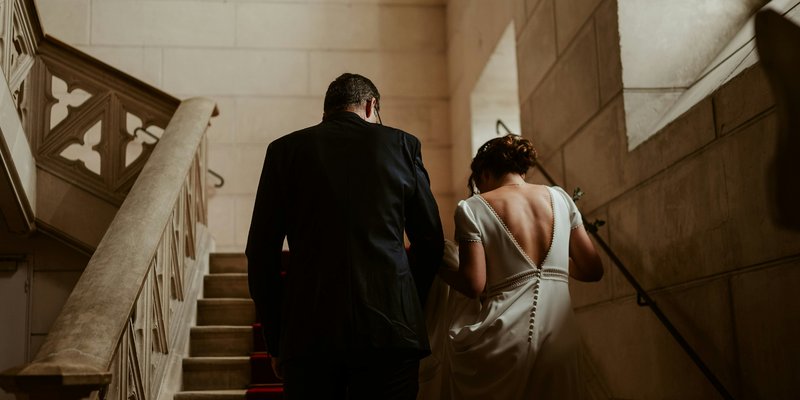 Central PA bride with an open back, short-sleeved wedding dress with lace detail and button going down the skirt walking with their backs to the camera up stone stairs with a groom in a dark suit.