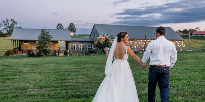 Bride and groom walk hand in hand across a grassy field at Honey Suckle Ridge, with the rustic barn venue glowing behind them under string lights at sunset.