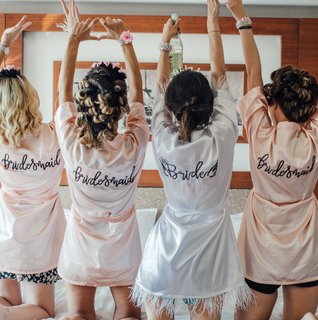 Four women wearing matching satin robes — three in blush pink that say “Bridesmaid” and one in white that says “Bride” — kneel on a hotel bed with their arms raised, celebrating together before the bachelorette festivities.