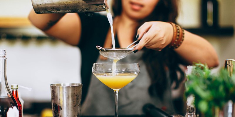 Bartender pours a freshly shaken yellow cocktail through a strainer into a coupe glass, surrounded by bar tools, bottles, and fresh ingredients.