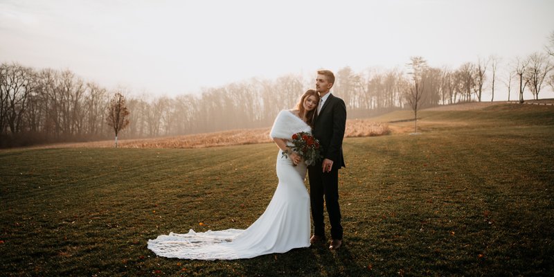 Bride and groom stand together in an open field at sunset, with the bride in a fitted white gown and faux fur wrap holding a bouquet of red roses, captured in a warm, moody style by Téa Ceresini Photography.
