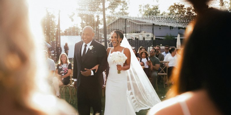 Bride walks down the aisle with her father during an outdoor wedding ceremony, smiling joyfully as sunlight filters through string lights and guests look on.