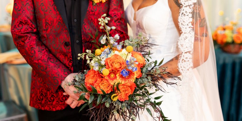 Bride and groom standing together holding a vibrant bouquet of orange, yellow, and blue flowers. The groom wears a red patterned jacket with a black shirt, while the bride wears a white gown with lace sleeves and a long veil.