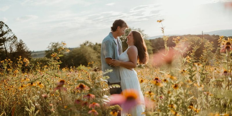 A couple shares a joyful moment surrounded by golden wildflowers during their engagement photo session at sunset in Central Pennsylvania, capturing a warm and romantic summer atmosphere.