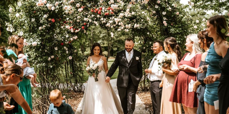 Bride and groom walk hand-in-hand beneath a floral archway surrounded by family and friends during their intimate outdoor wedding ceremony at Moonstone Manor in Central Pennsylvania.