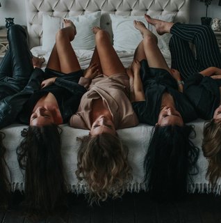 Five women lie on a bed, heads upside down, in a cozy room. Plants and soft lighting enhance the relaxed, playful atmosphere.