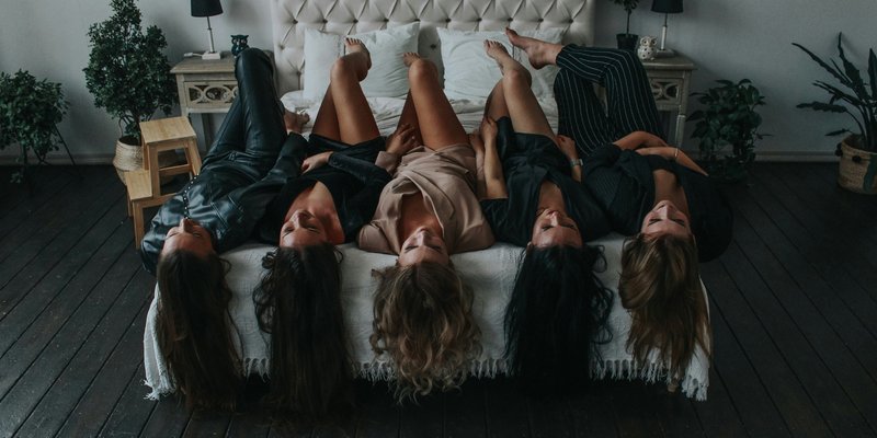 Five women lie on a bed, heads upside down, in a cozy room. Plants and soft lighting enhance the relaxed, playful atmosphere.
