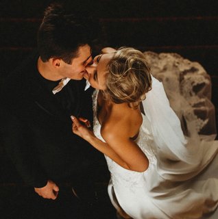 Overhead view of a bride and groom kissing on a staircase, with the bride in a white lace gown and veil and the groom in a black suit during their wedding.