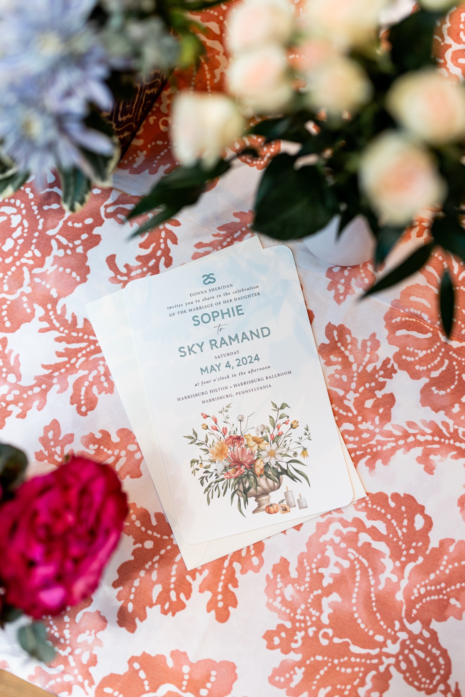 Wedding invitation on a coral-patterned tablecloth, surrounded by blue and pink flowers. The card features floral illustration and says "May 4, 2024."