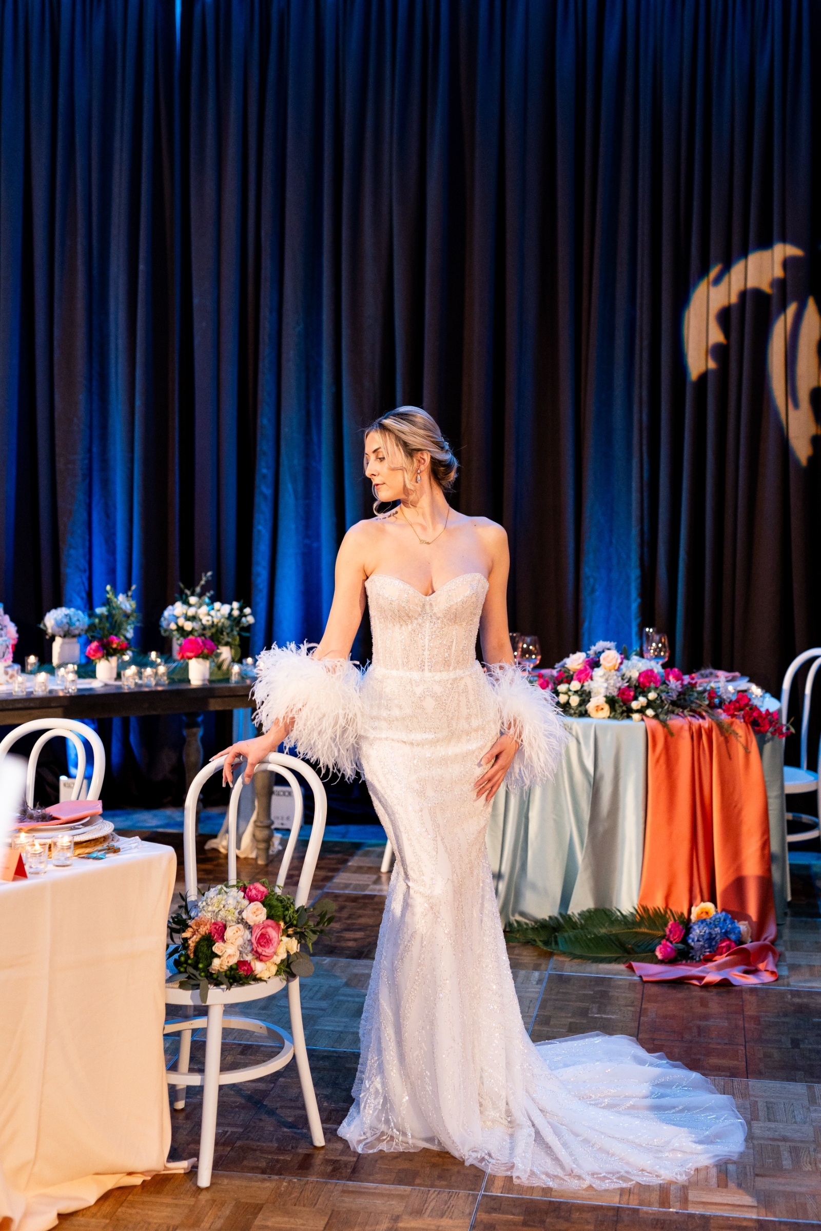 Bride posing beside a colorful wedding tablescape featuring coral satin, pastel florals, and romantic candlelight.