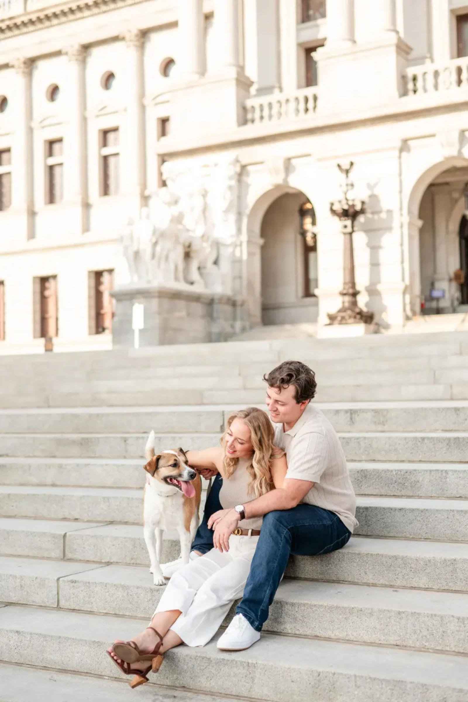Couple sitting on the Pennsylvania State Capitol steps with their dog, smiling during their relaxed and playful engagement photos in Harrisburg.