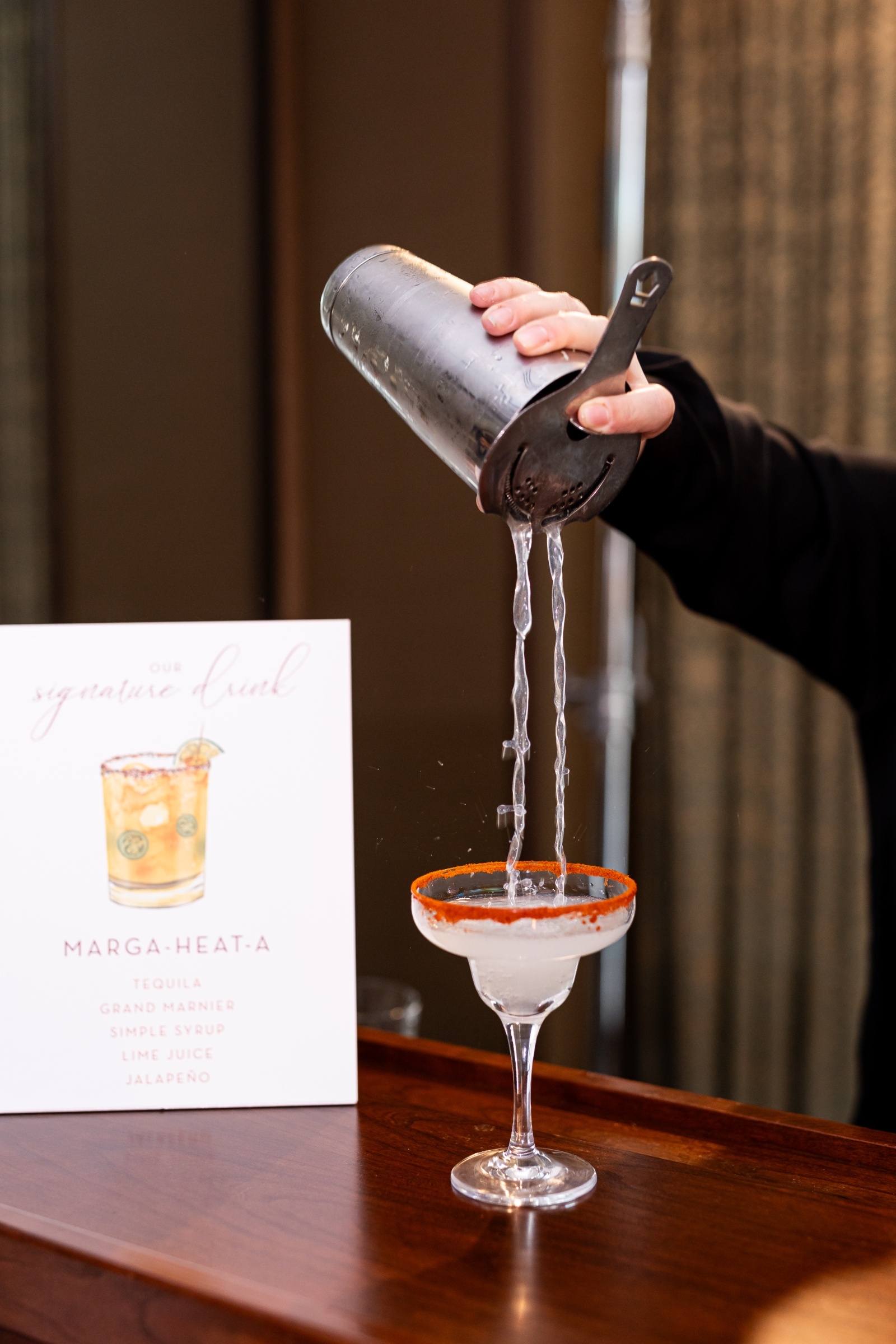 Bartender pouring a margarita into a glass with a chili-salt rim beside a cocktail sign reading “Marga-Heat-A,” featuring tequila, lime, and jalapeño.