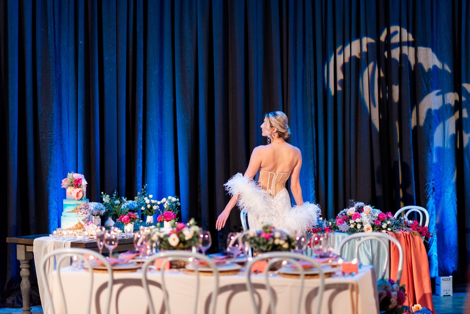 Bride in a glamorous strapless gown with feathered sleeves walking through a Mediterranean-inspired wedding reception with blue and coral details.