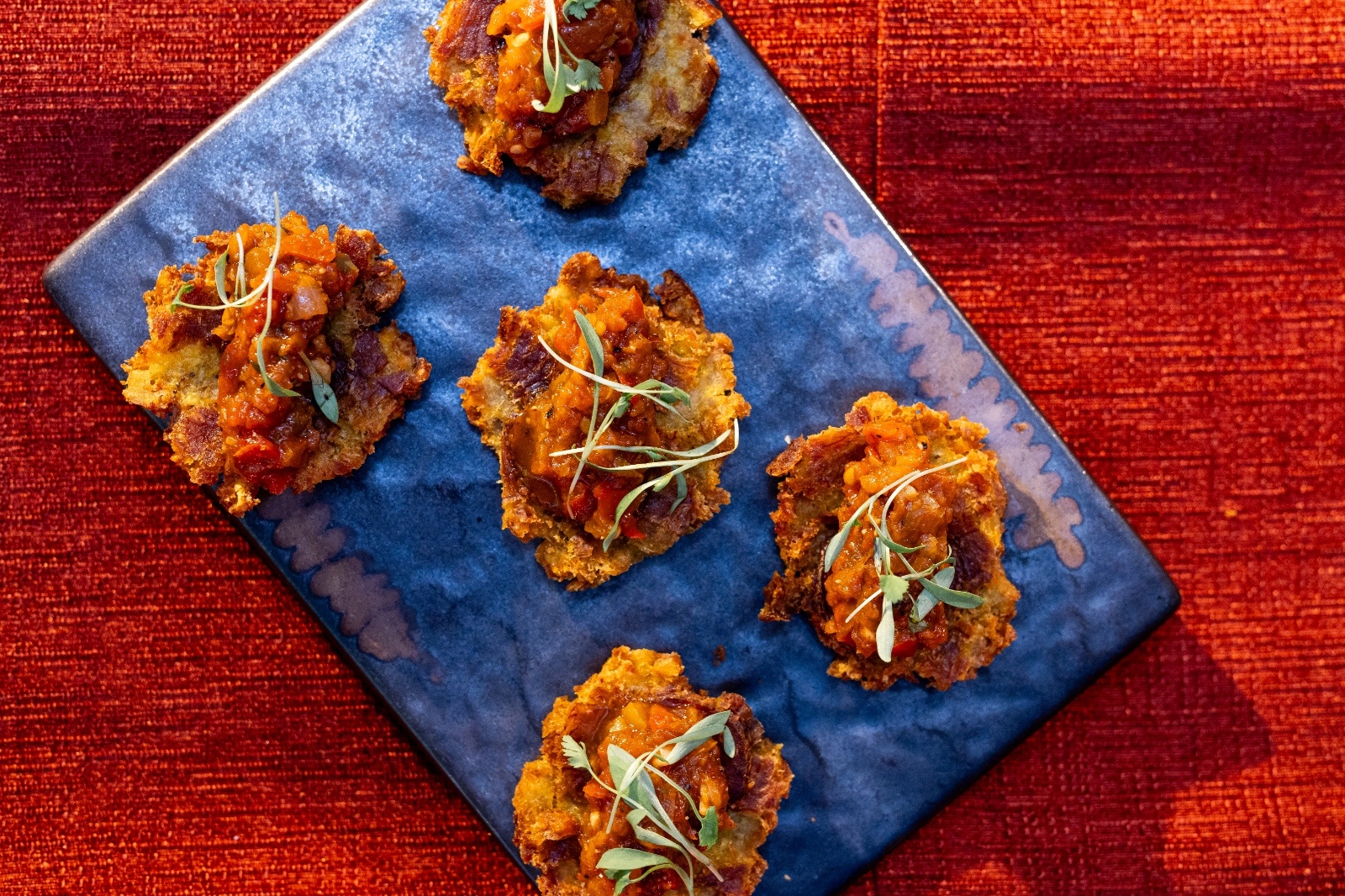 Close-up of bite-sized appetizers topped with tomato chutney and microgreens, arranged on a blue plate over a rich red tablecloth.
