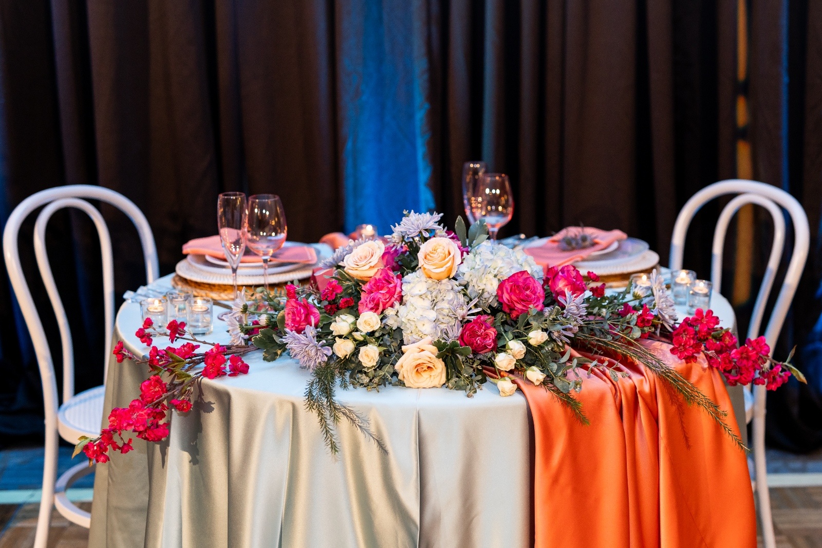 Elegant round wedding table with turquoise satin linens, coral runner, and a lush floral centerpiece featuring pink roses, cream blooms, and hydrangeas.