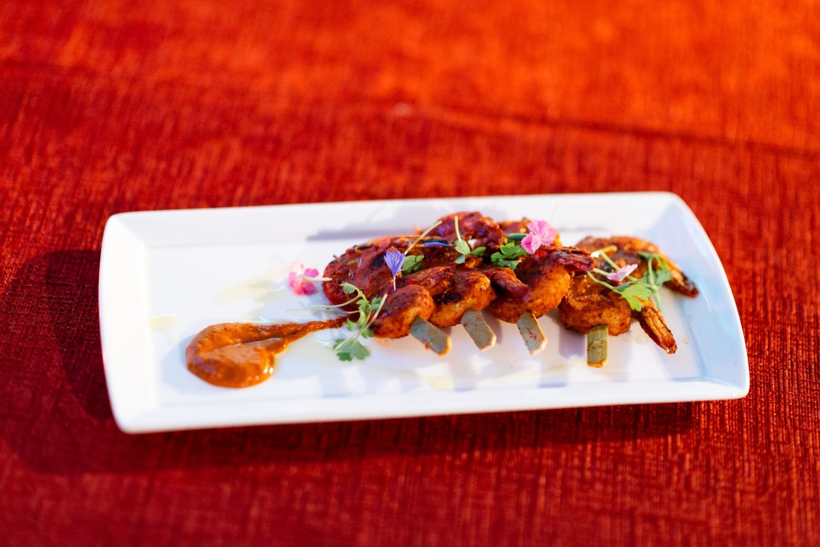 Plated shrimp skewers garnished with edible flowers and herbs, served with a spicy dipping sauce on a white rectangular dish.