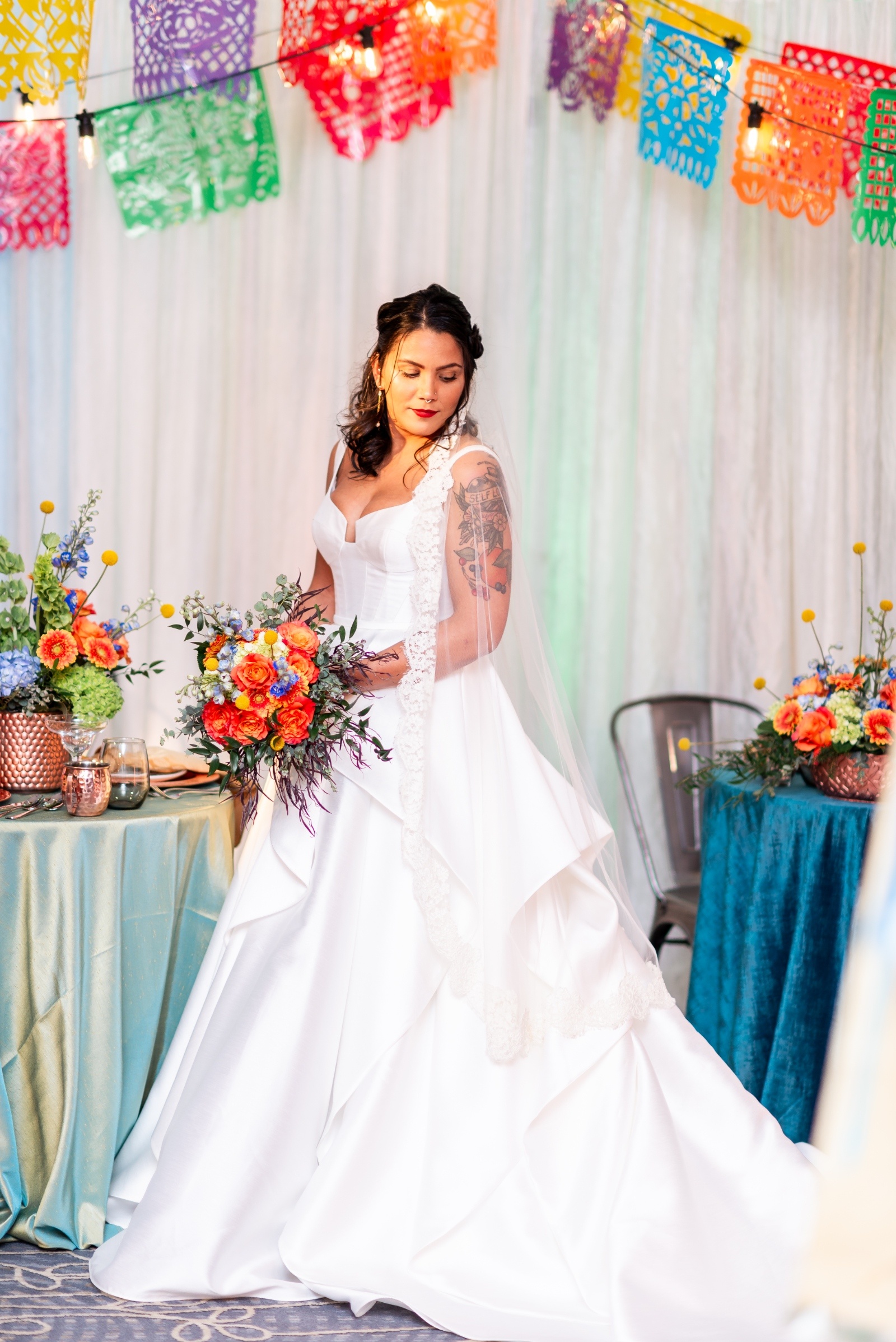 Bride in a white satin gown holding a colorful bouquet beneath papel picado banners, surrounded by vibrant floral table décor inspired by Latin wedding traditions.