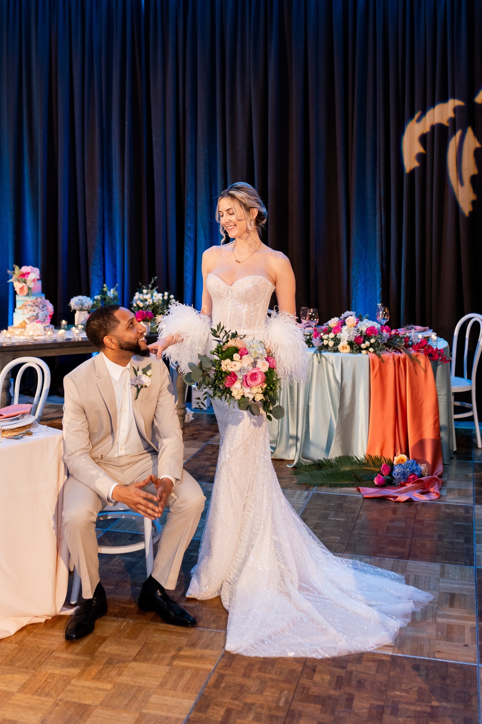 Bride in a strapless beaded gown with feathered sleeves smiling at the groom in a tan suit during a Mediterranean-inspired wedding reception.