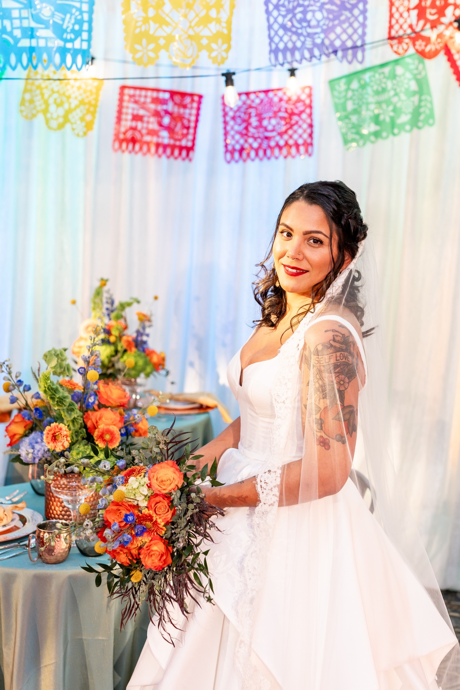Bride in a modern satin gown and lace veil smiling while holding a vivid bouquet of orange and blue blooms under festive papel picado banners.