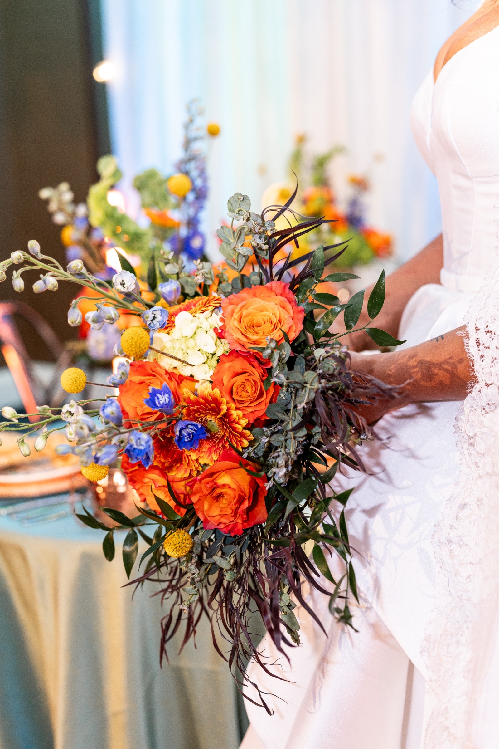 Close-up of a wedding bouquet featuring Verona sunset orange roses, blue delphinium, yellow billy balls, and greenery, captured against teal table linens.