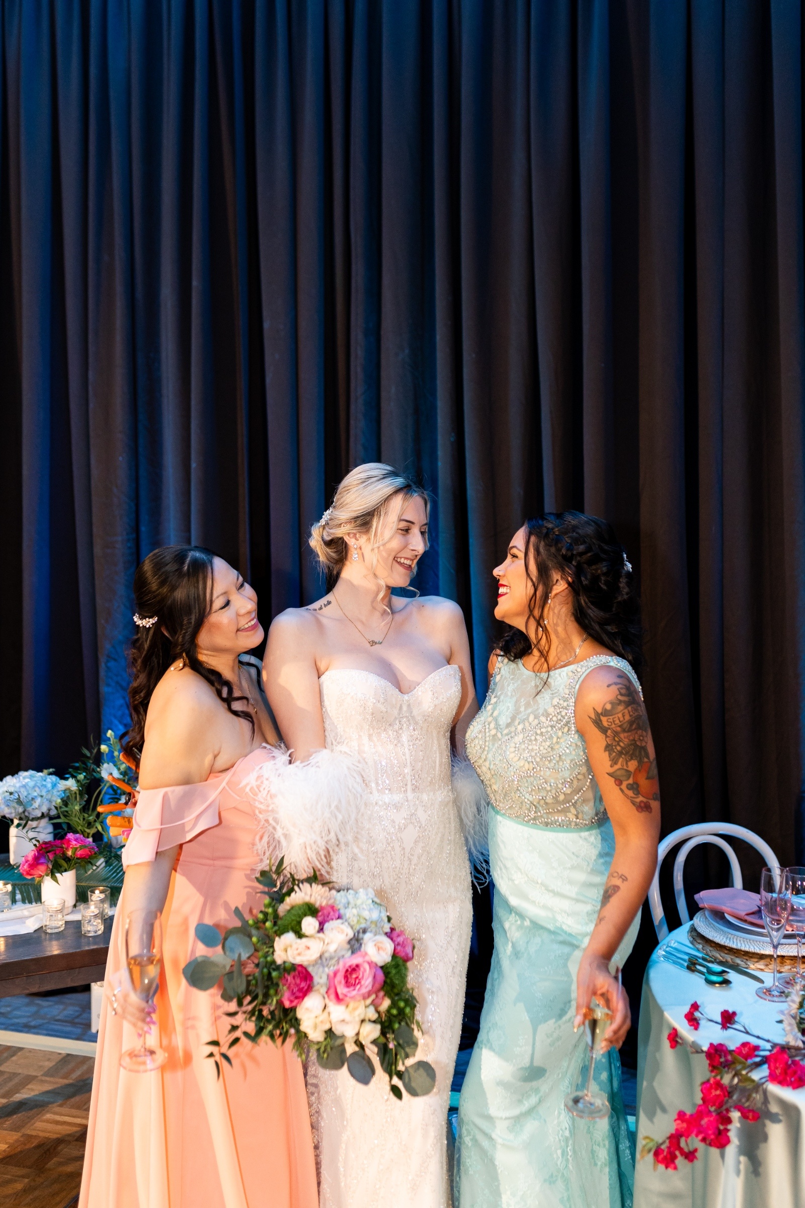 Bride and bridesmaids sharing a joyful moment together, dressed in blush and aqua gowns, with vibrant floral bouquets.