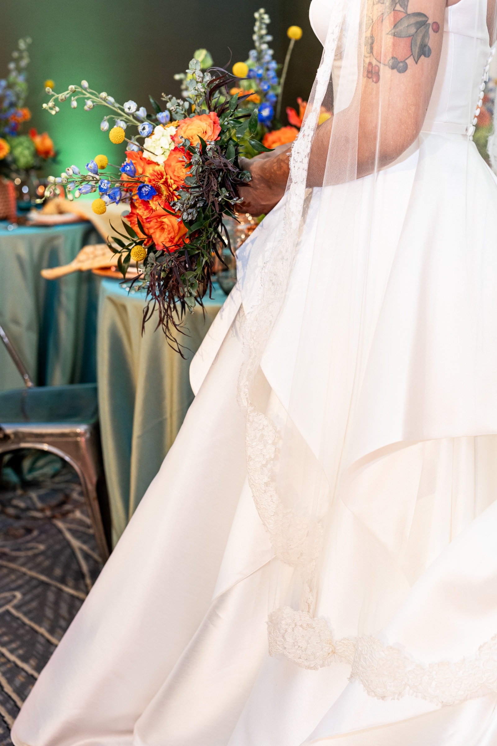 Bride’s white gown and lace veil draped over her arm as she holds a cascading bouquet of orange, blue, and yellow florals.