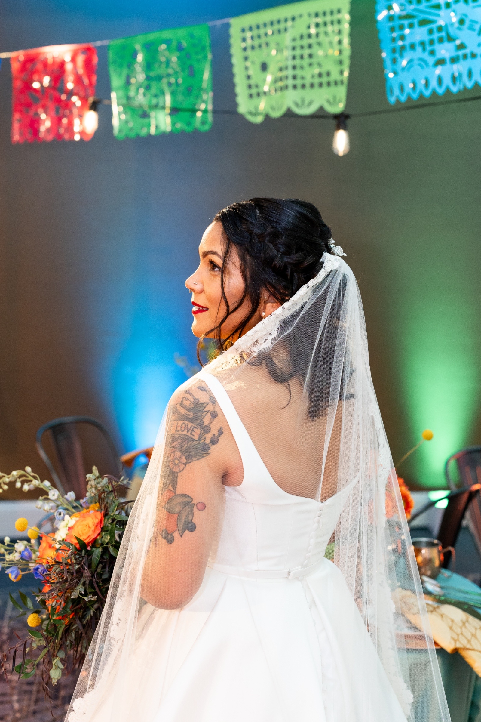 Bride with a braided hairstyle and lace veil glancing over her shoulder beneath colorful papel picado banners at a festive wedding reception.