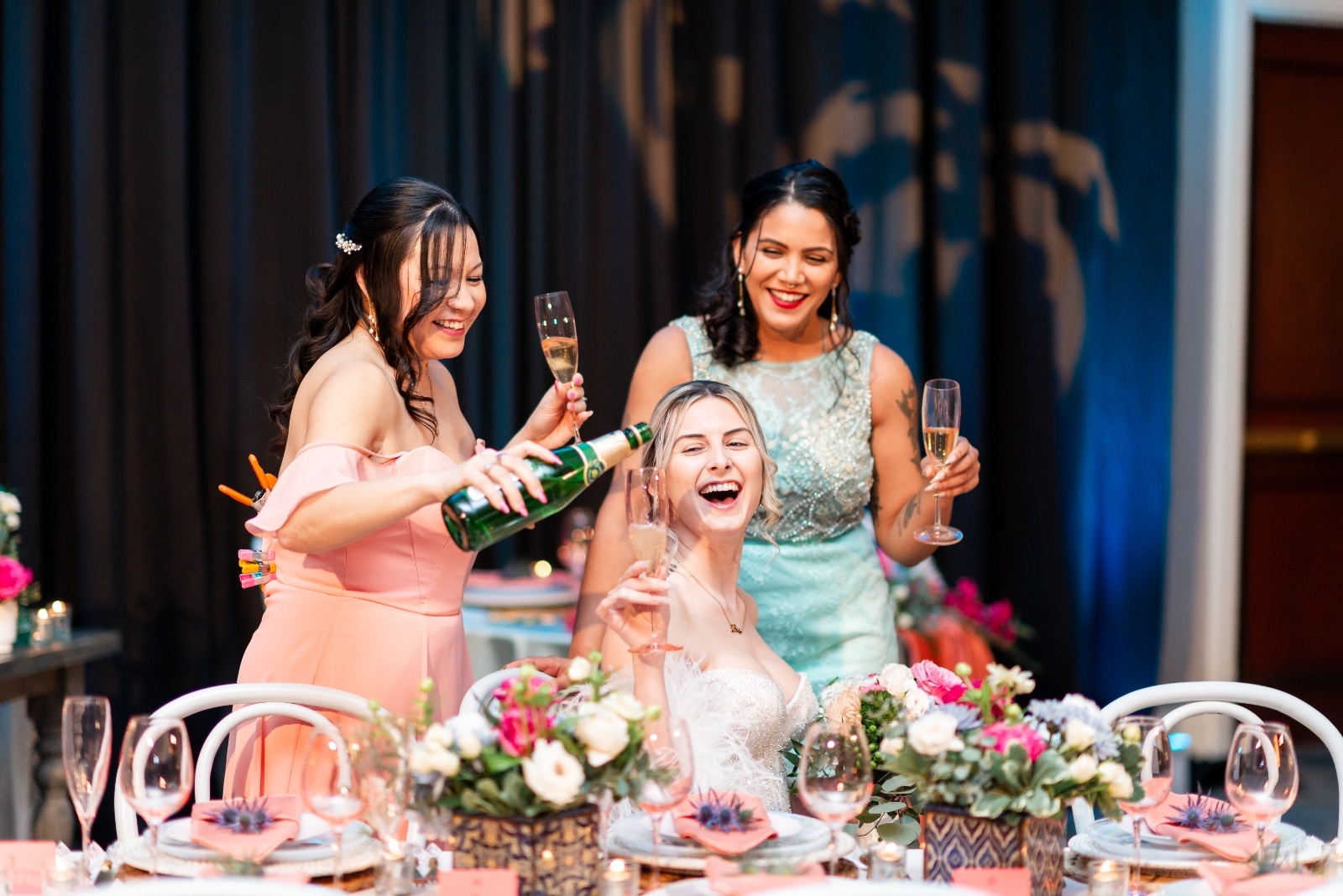 Bride laughing as bridesmaids pour champagne during a colorful wedding celebration with coral and turquoise decor.