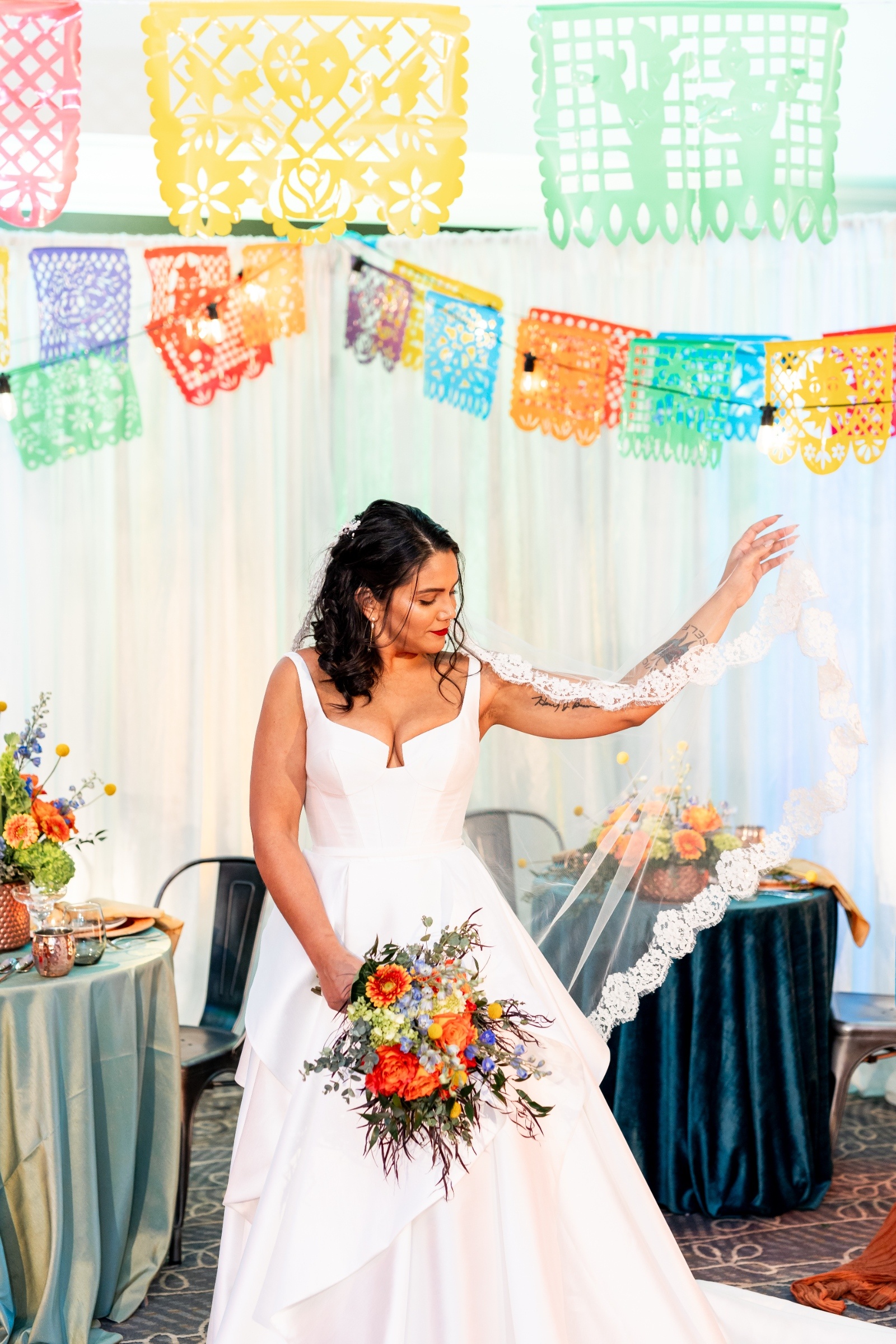 Bride in a modern white dress gracefully holding her lace veil while displaying a bright bouquet of orange and blue flowers under papel picado décor.