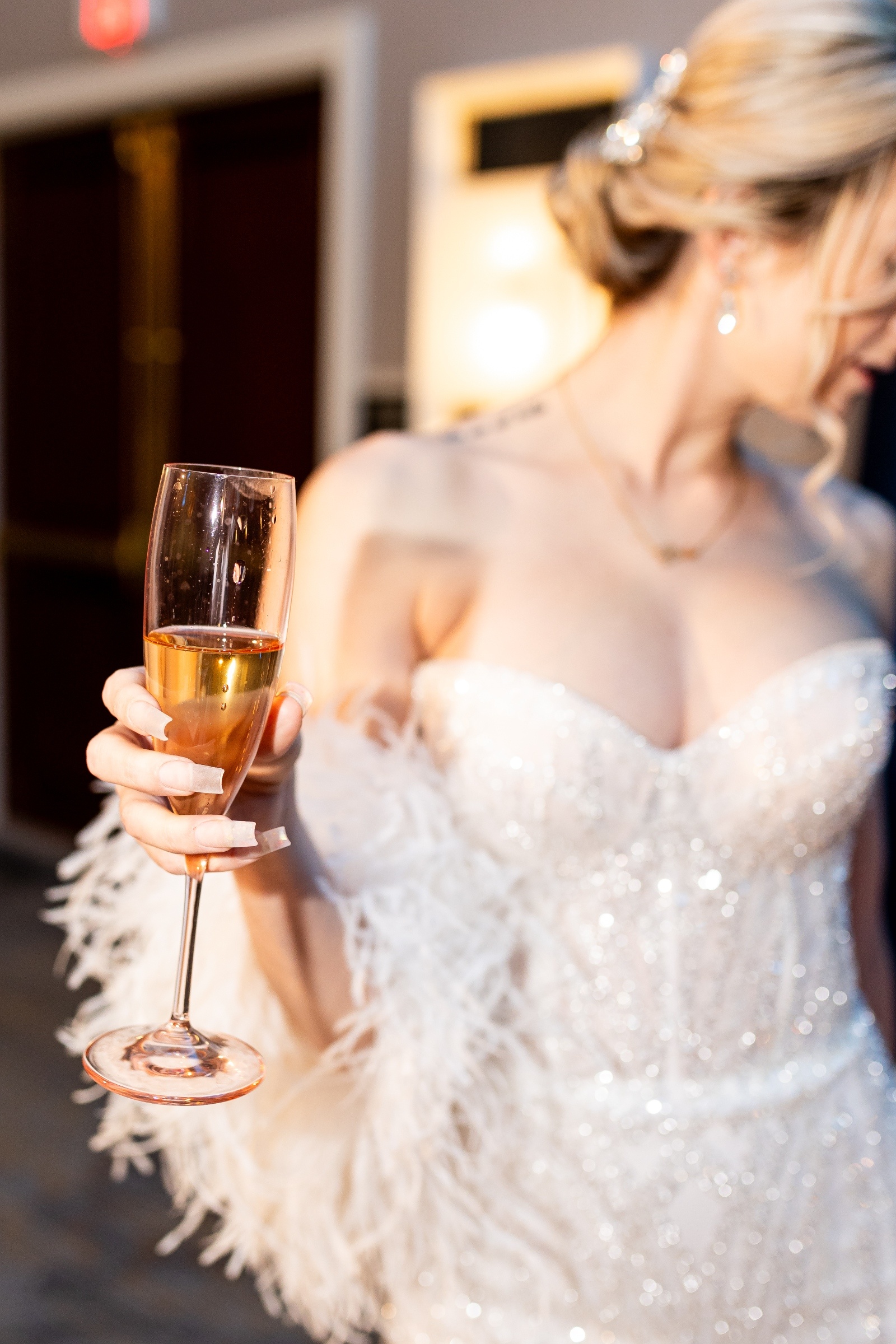 Bride in a sparkling strapless gown holding a champagne flute, showcasing soft glam bridal style with feathered details