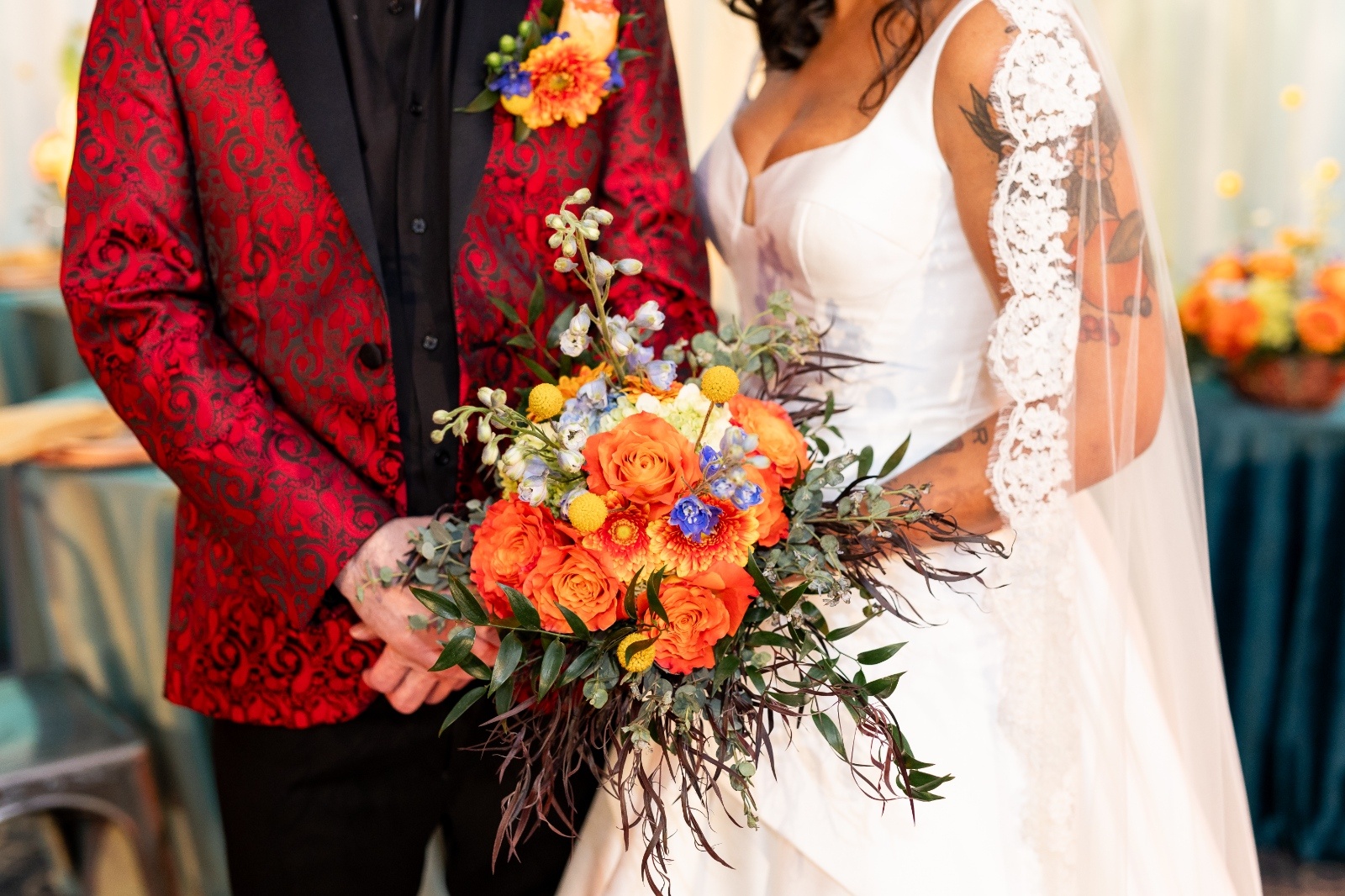 Bride and groom standing side by side with a bold bouquet of verona sunset orange roses and blue florals against colorful Latin-inspired wedding décor.