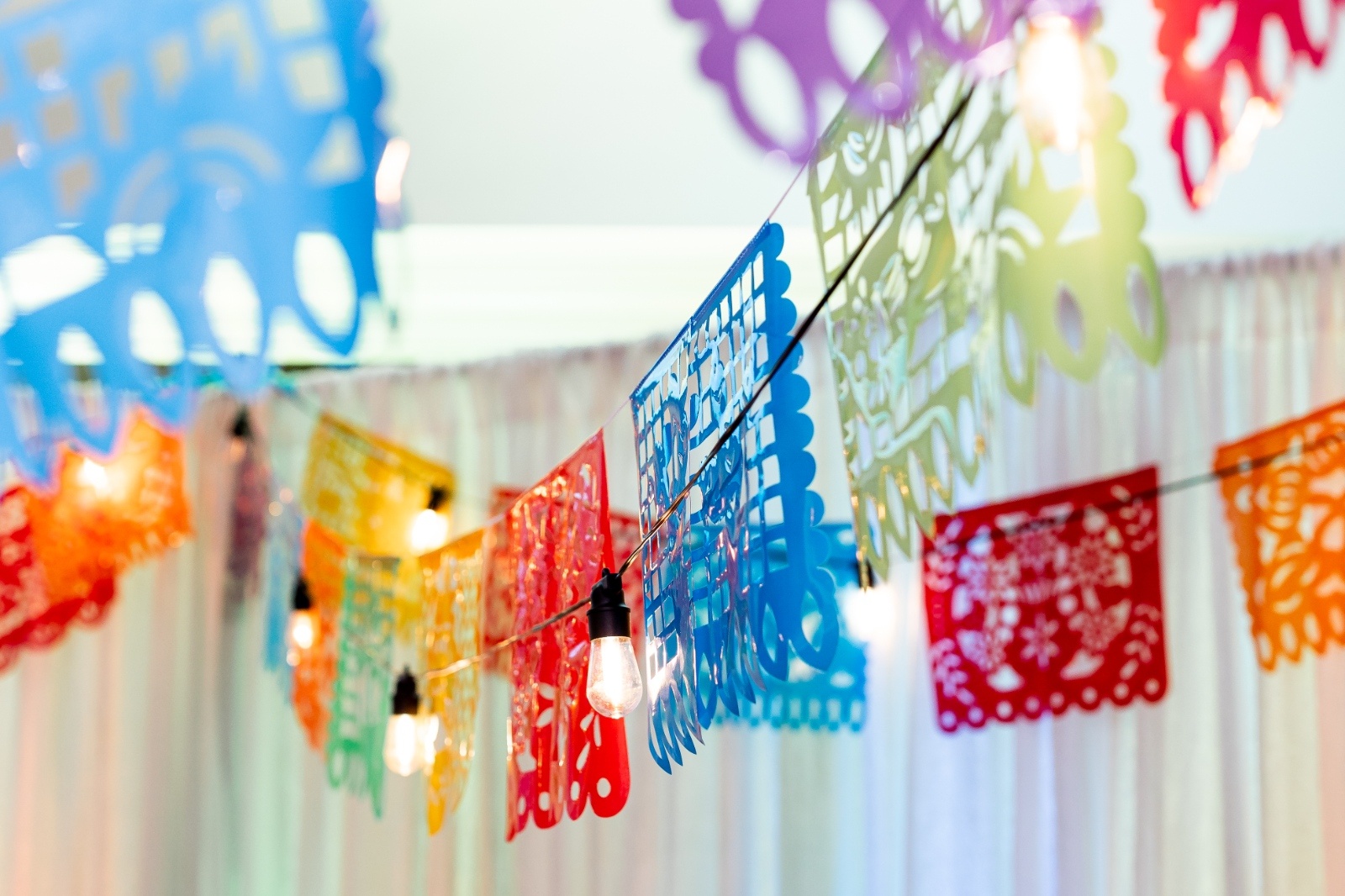 Brightly colored papel picado banners in red, blue, yellow, green, and purple hang across a reception space, illuminated by string lights for a festive wedding atmosphere.