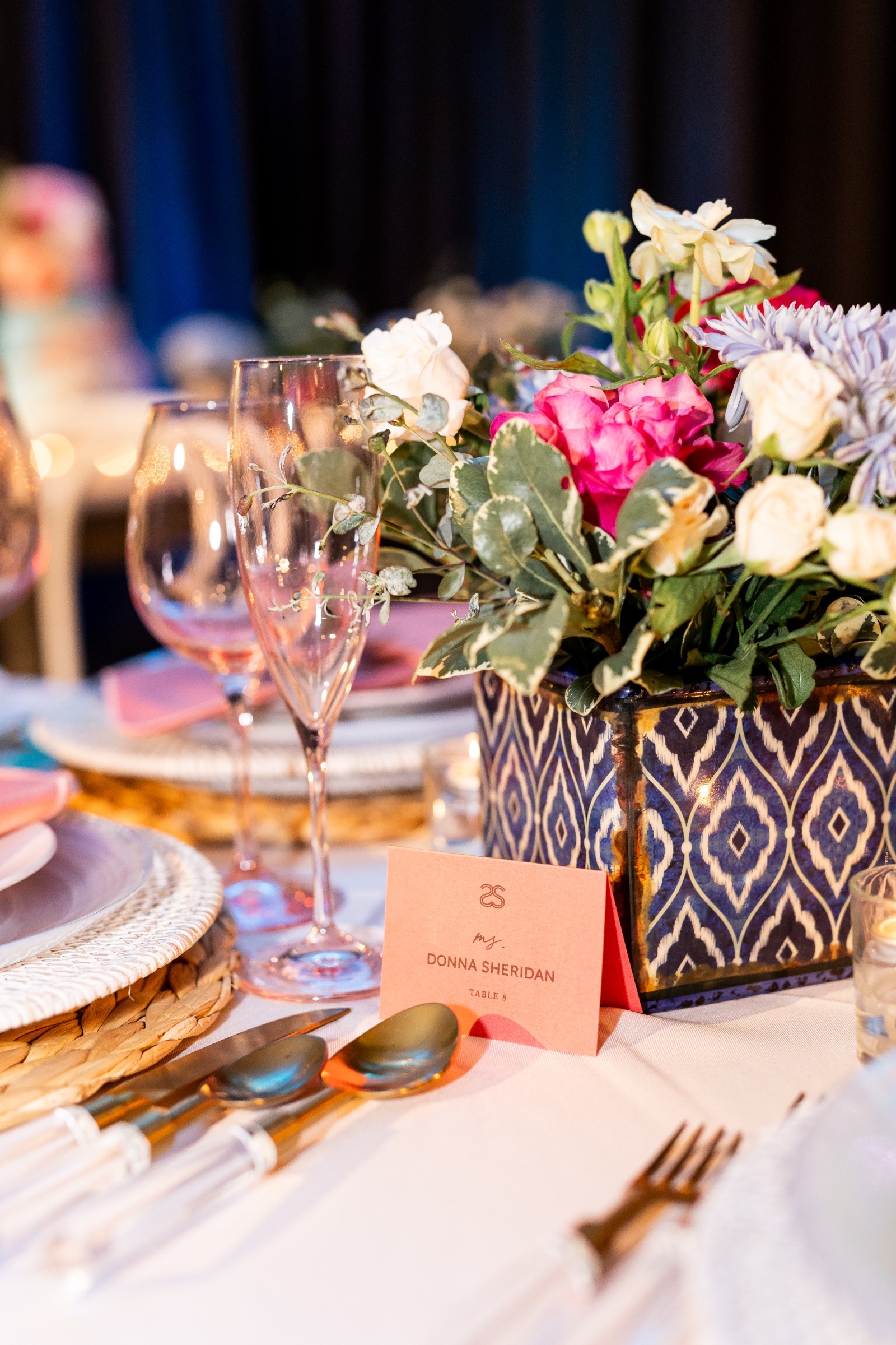 Elegant table setting with a blue patterned vase holding white, pink, and purple flowers. Two champagne flutes and a place card labeled "Donna Sheridan" adorn the table.