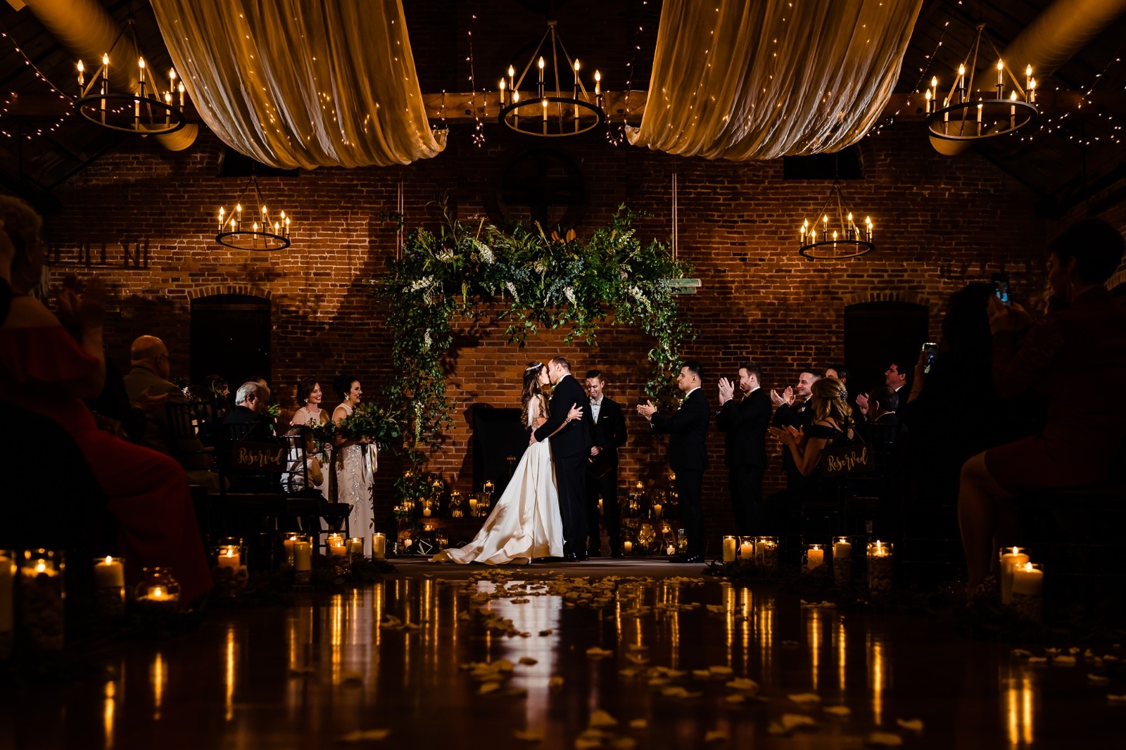 Bride and groom kissing at the end of a candlelit aisle under lush greenery.