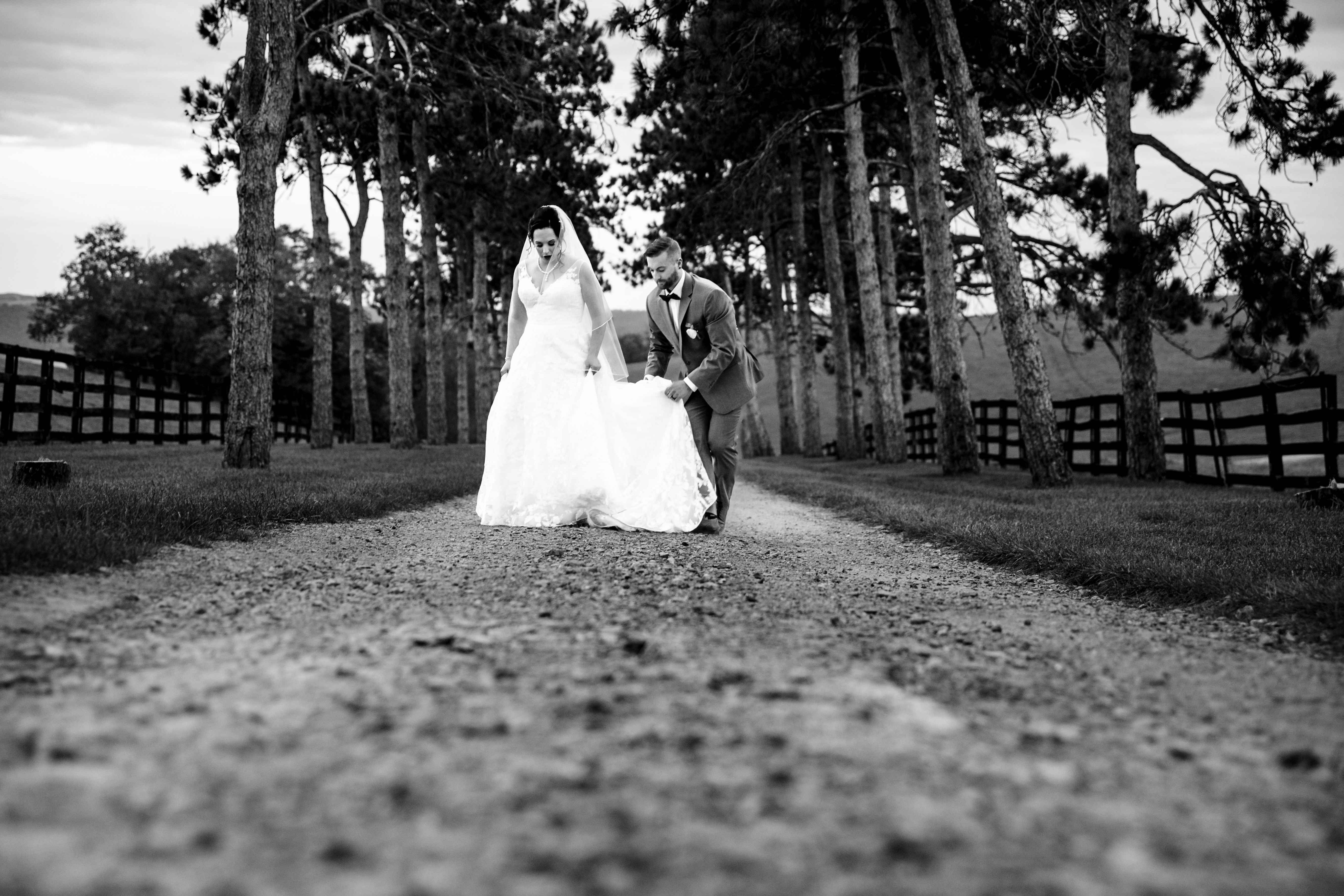 Groom helps adjust the bride's dress as they walk down a tree-lined gravel lane at a Pennsylvania wedding venue.