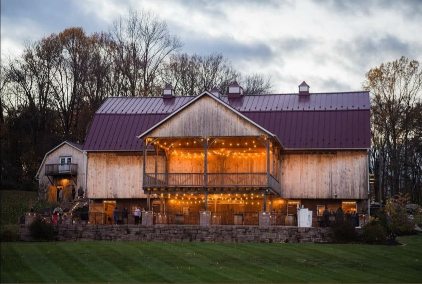 The barn at Spring Valley Farms glows with warm string lights at dusk, showcasing its wooden exterior and countryside charm surrounded by trees and open fields.