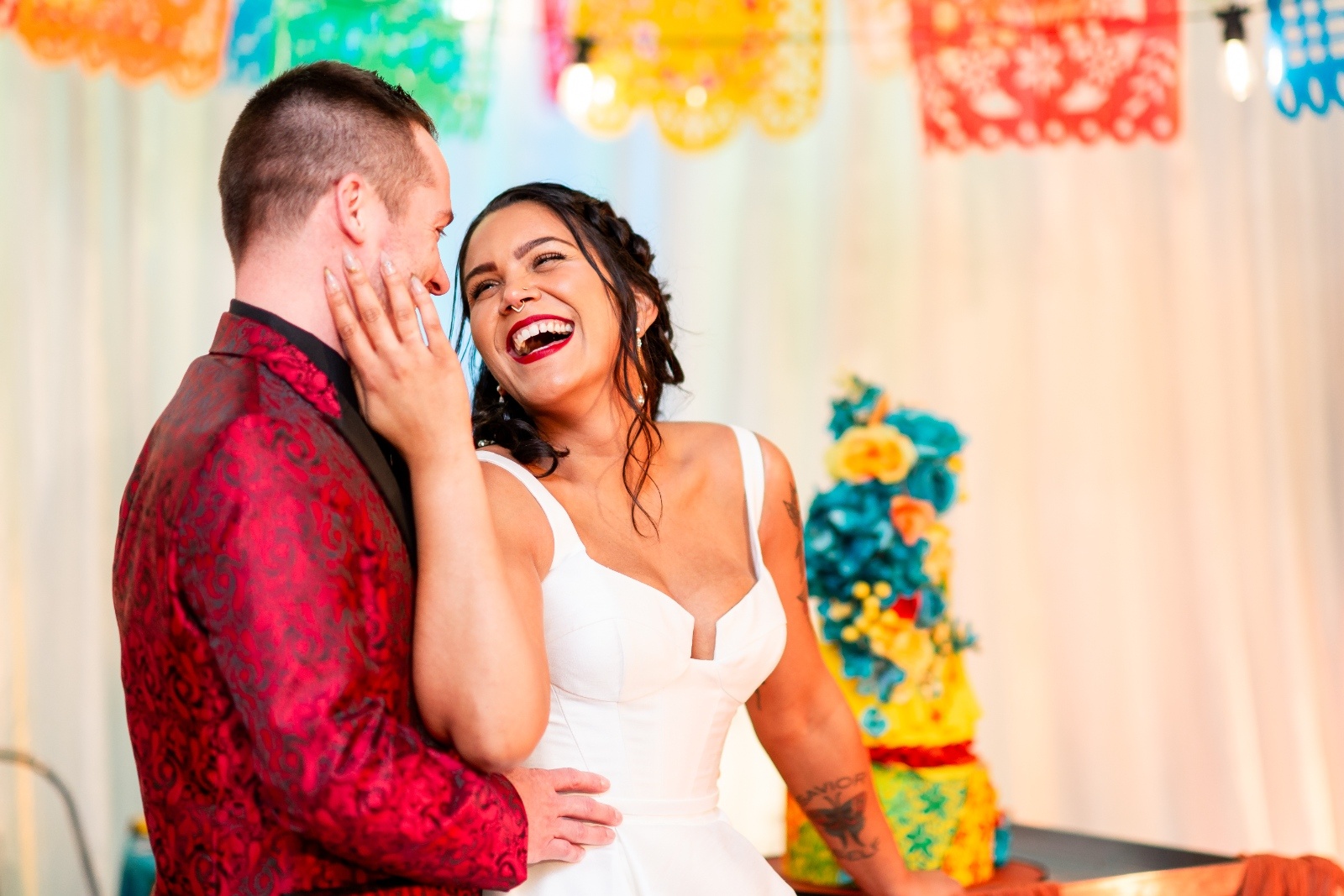 Joyful bride and groom sharing a laugh under colorful papel picado banners with a bright floral cake in the background.