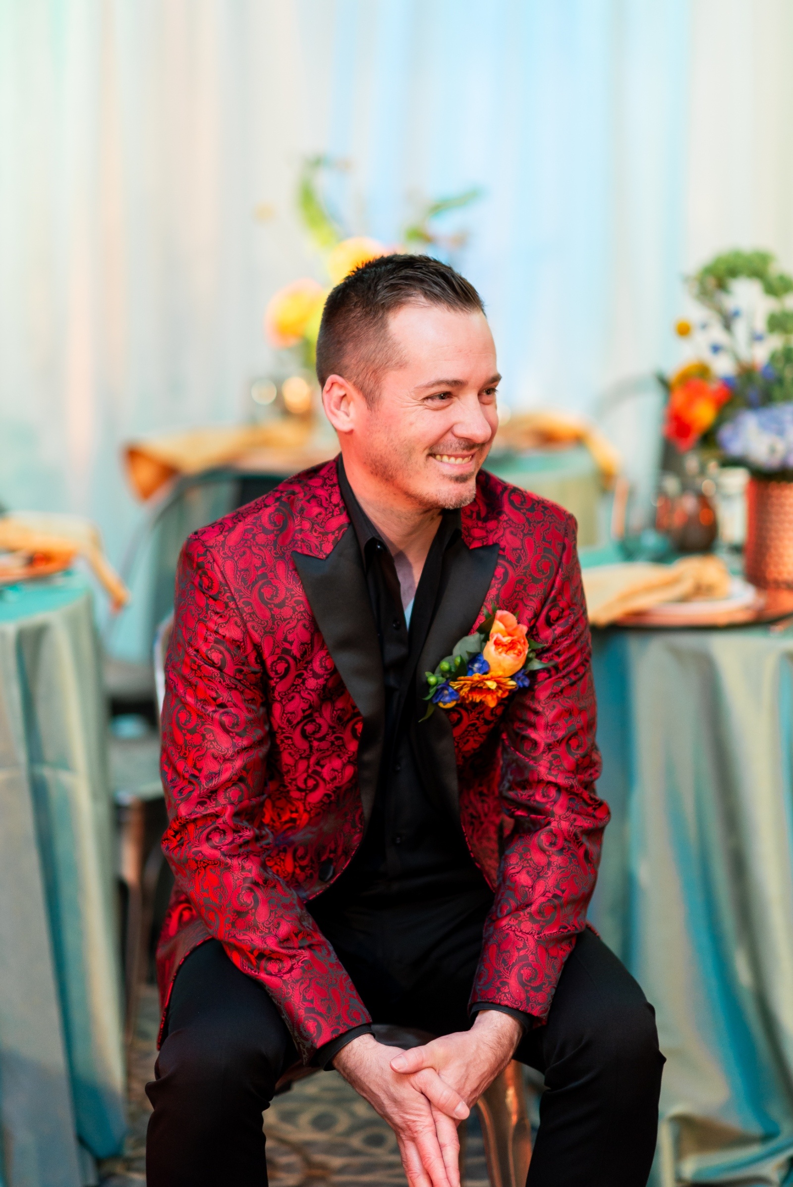 Groom smiling in a red paisley jacket accented with an orange and blue boutonniere, seated among colorful wedding table décor.