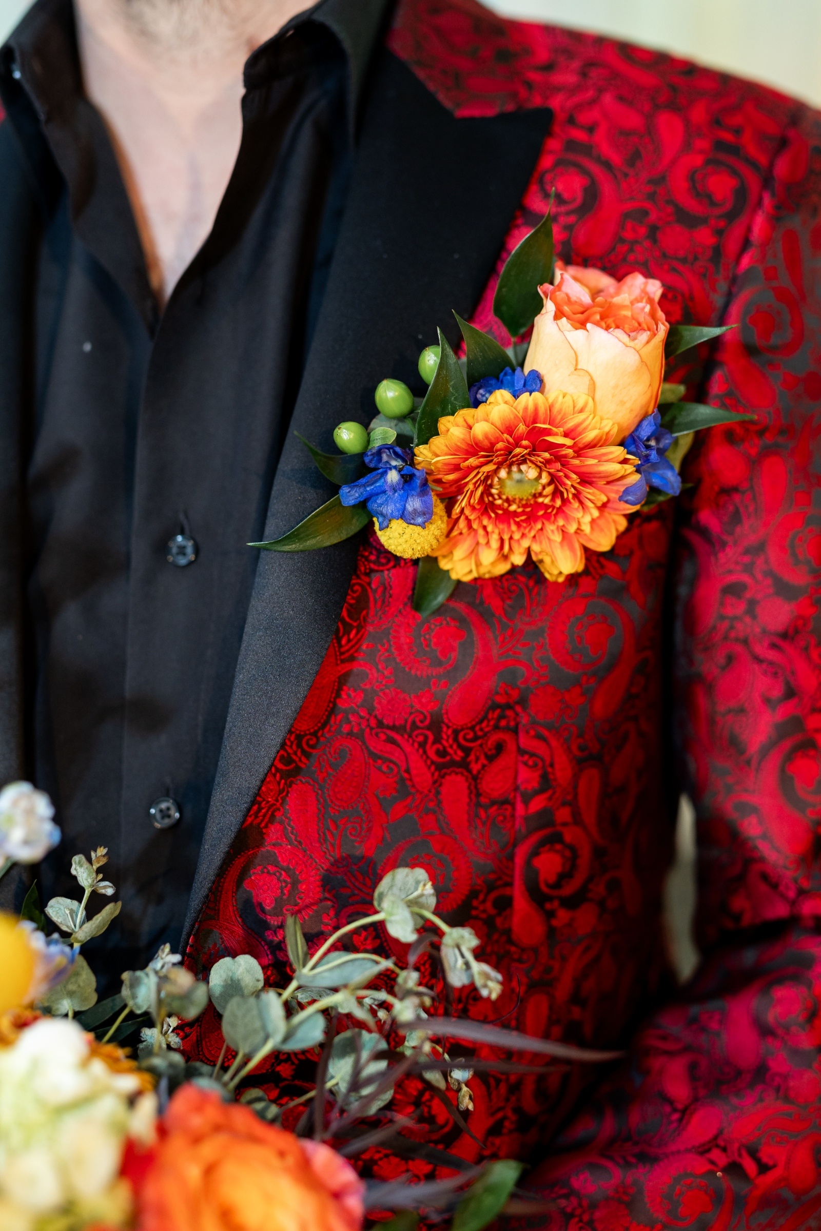 Close-up of a groom’s boutonniere with orange rose, blue delphinium, and yellow billy ball on a red paisley suit jacket.
