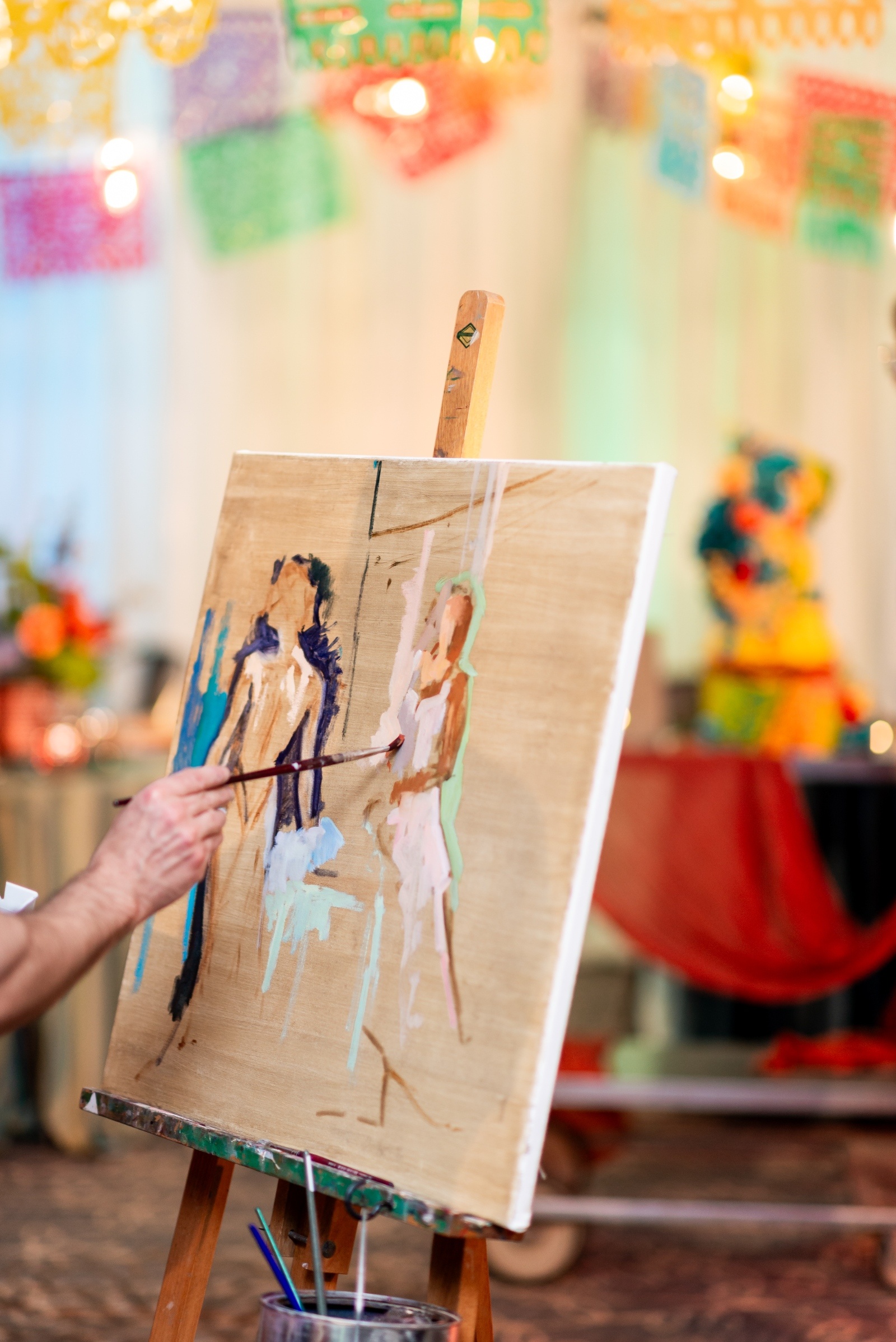 Artist painting a live wedding portrait of the couple on canvas, with colorful papel picado banners and a bright floral cake in the background.