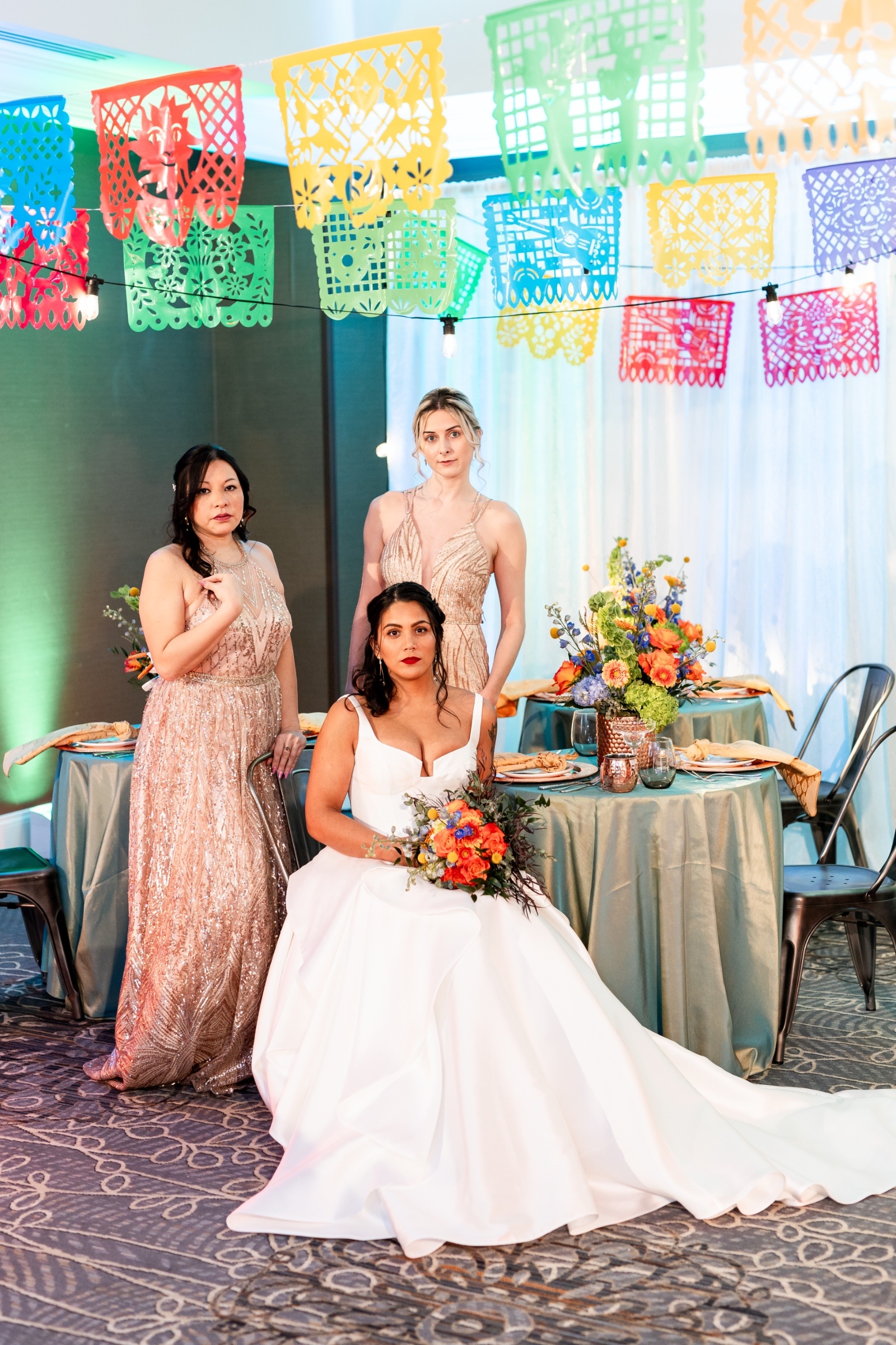 Bride in a white gown with two bridesmaids in gold dresses standing beneath colorful papel picado banners at a fiesta-inspired wedding reception.