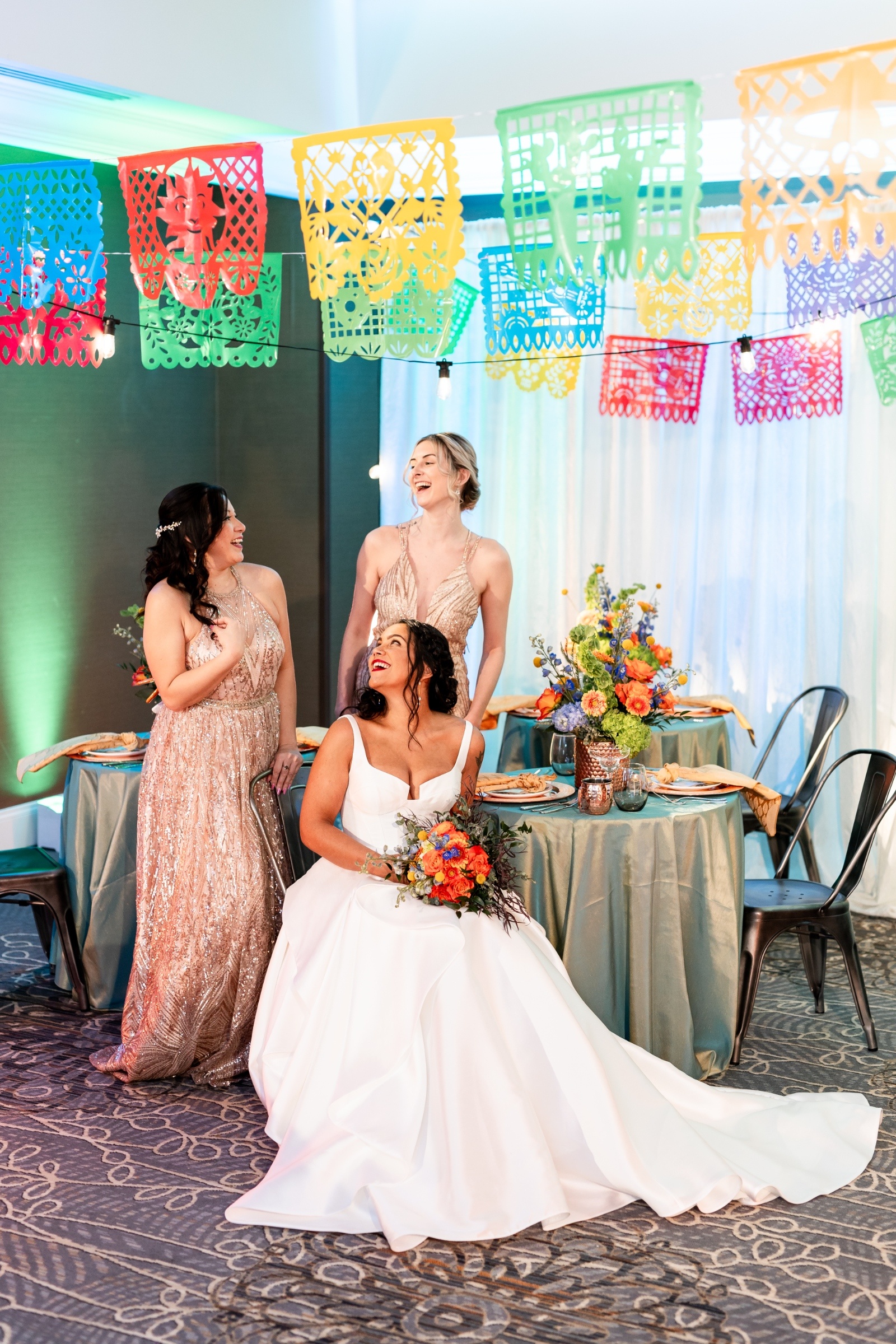 Bride and her bridesmaids sharing a joyful laugh under festive papel picado banners, surrounded by bright floral centerpieces and teal table linens.