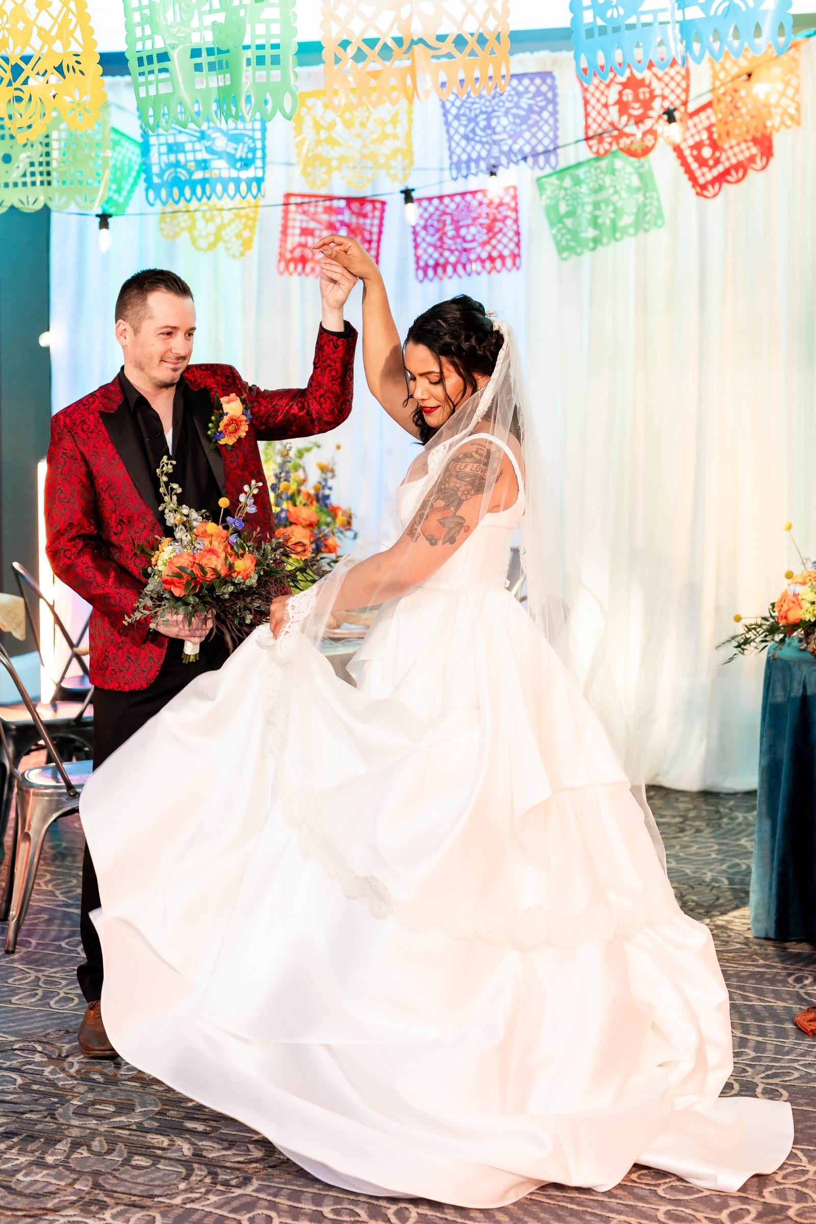 Groom in a red patterned jacket twirls the bride in her white gown during a lively dance under colorful papel picado wedding decorations.
