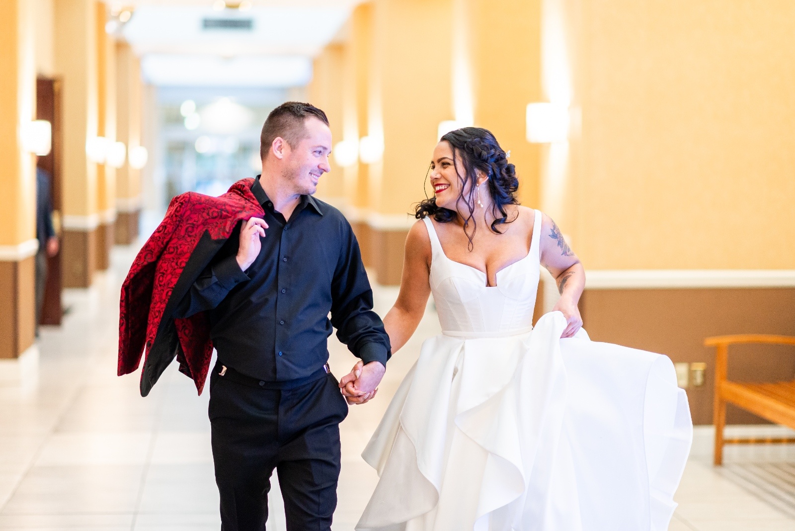 Bride in a white dress and groom in a black shirt with red jacket draped over his shoulder walking hand in hand through a bright hallway, smiling at each other.