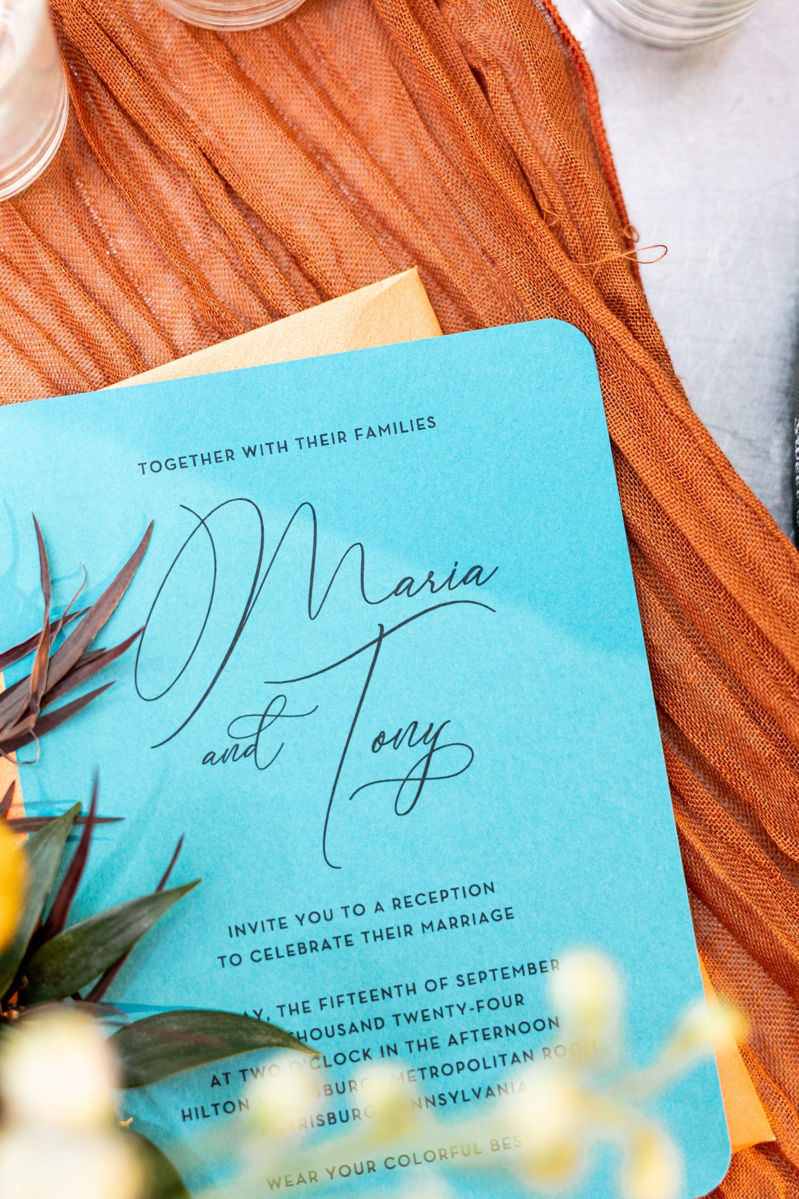 Close-up of a teal wedding invitation reading “Maria and Tony,” styled on a rust-colored fabric with delicate floral accents.