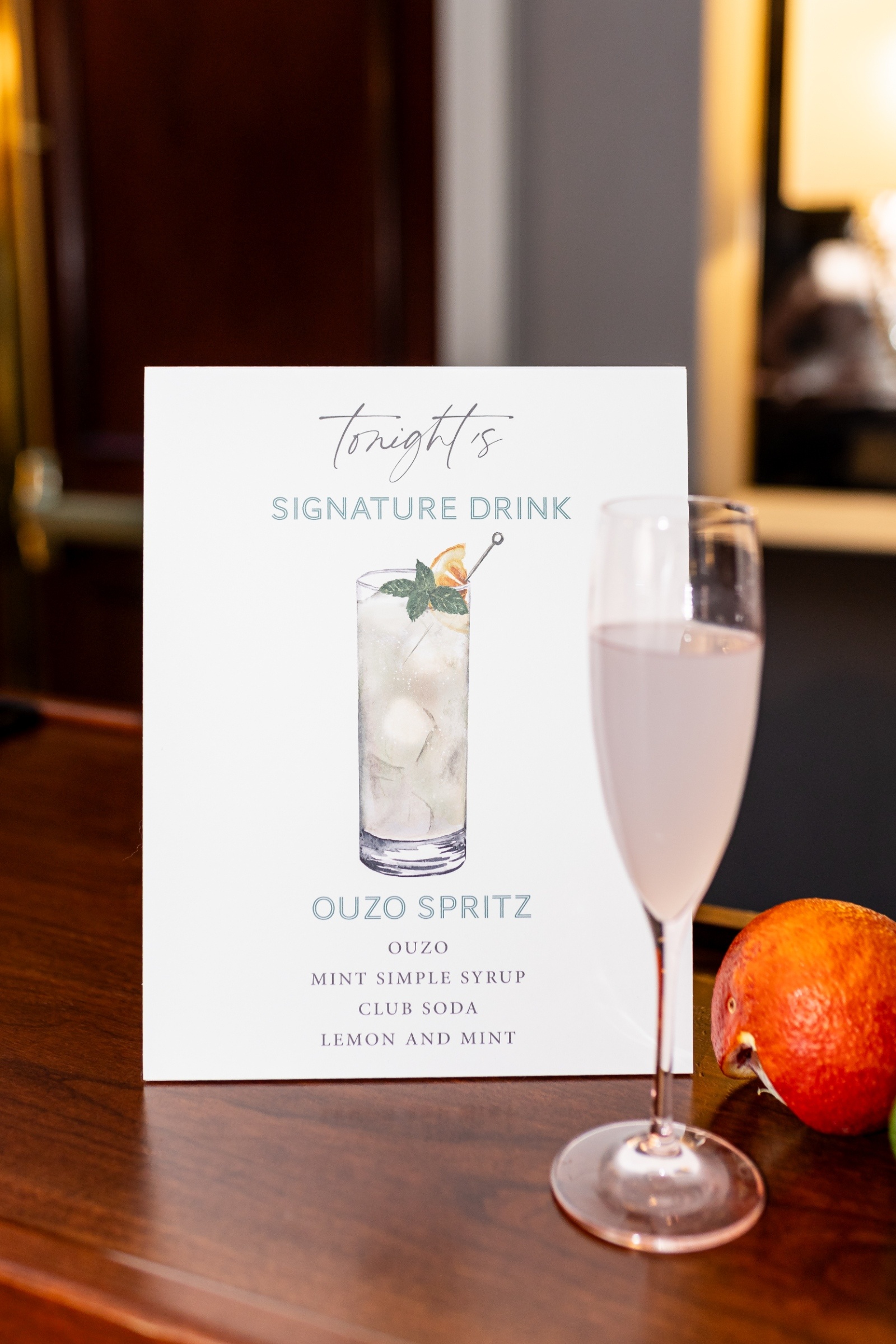 A menu card on a wooden bar features "Tonight's Signature Drink: Ouzo Spritz" with ingredients listed. A glass of the spritz and an orange are nearby.