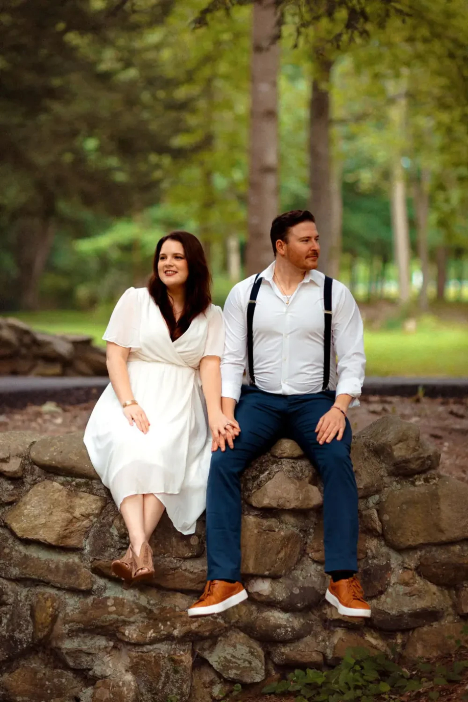 Couple holding hands while sitting on a rustic stone wall surrounded by tall evergreens during their engagement session in Caledonia State Park, Fayetteville, Pennsylvania.
