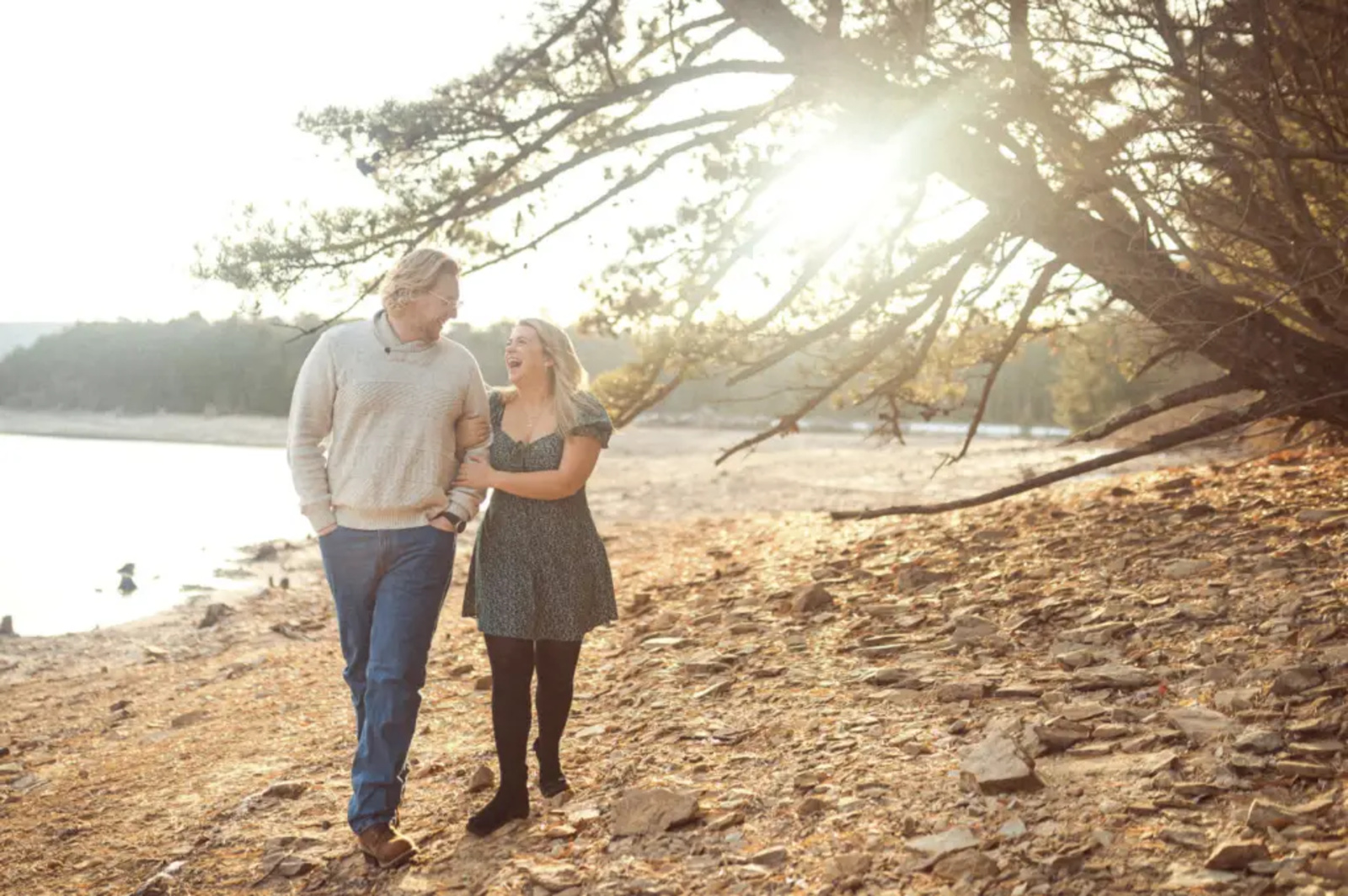 Couple walking arm-in-arm along the rocky shoreline of Long Pine Run Reservoir in Caledonia State Park, laughing together as the evening sun filters through the trees in Fayetteville, PA.