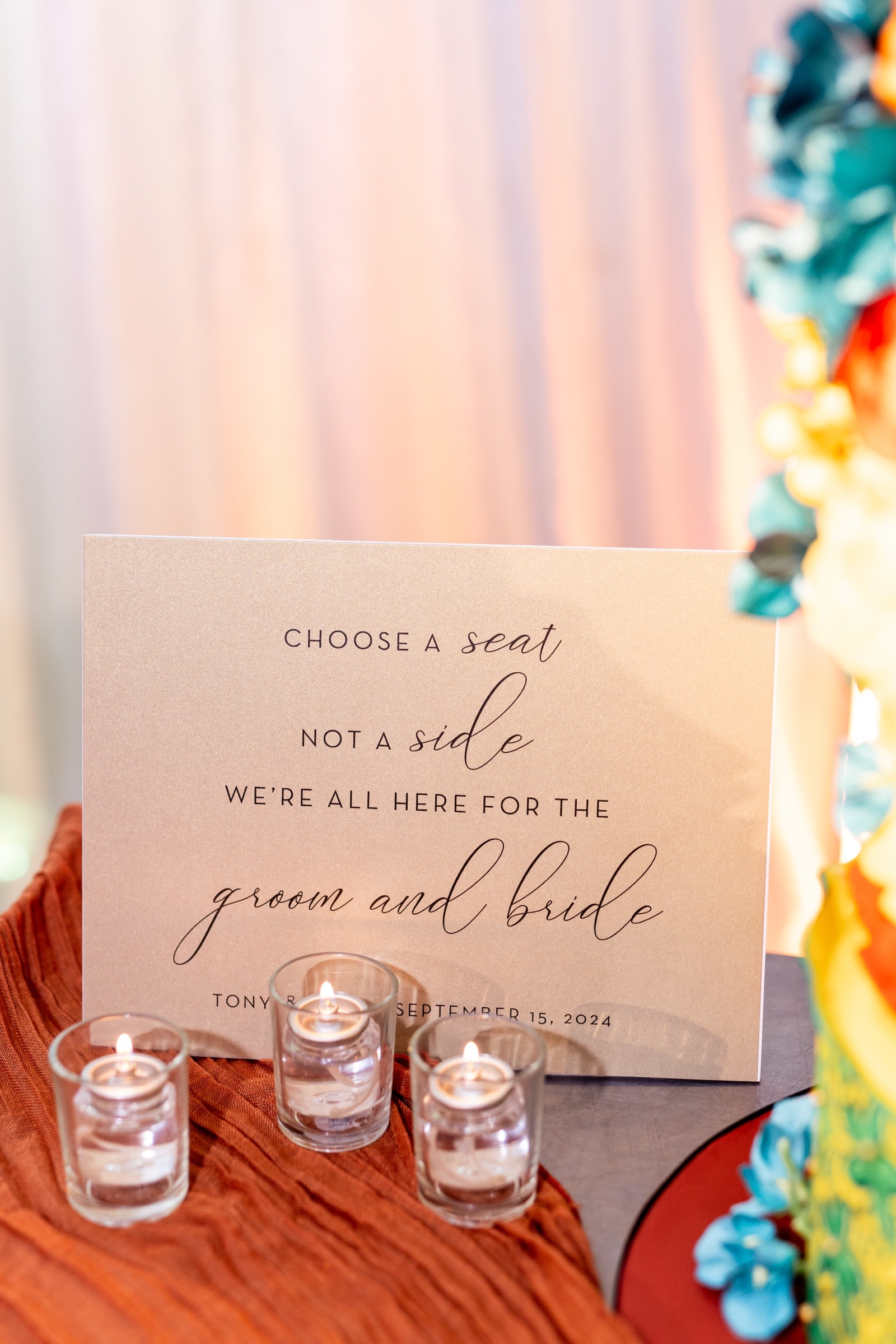 Beige wedding sign reading “Choose a seat, not a side, we’re all here for the groom and bride,” displayed with glowing votive candles and warm tones.
