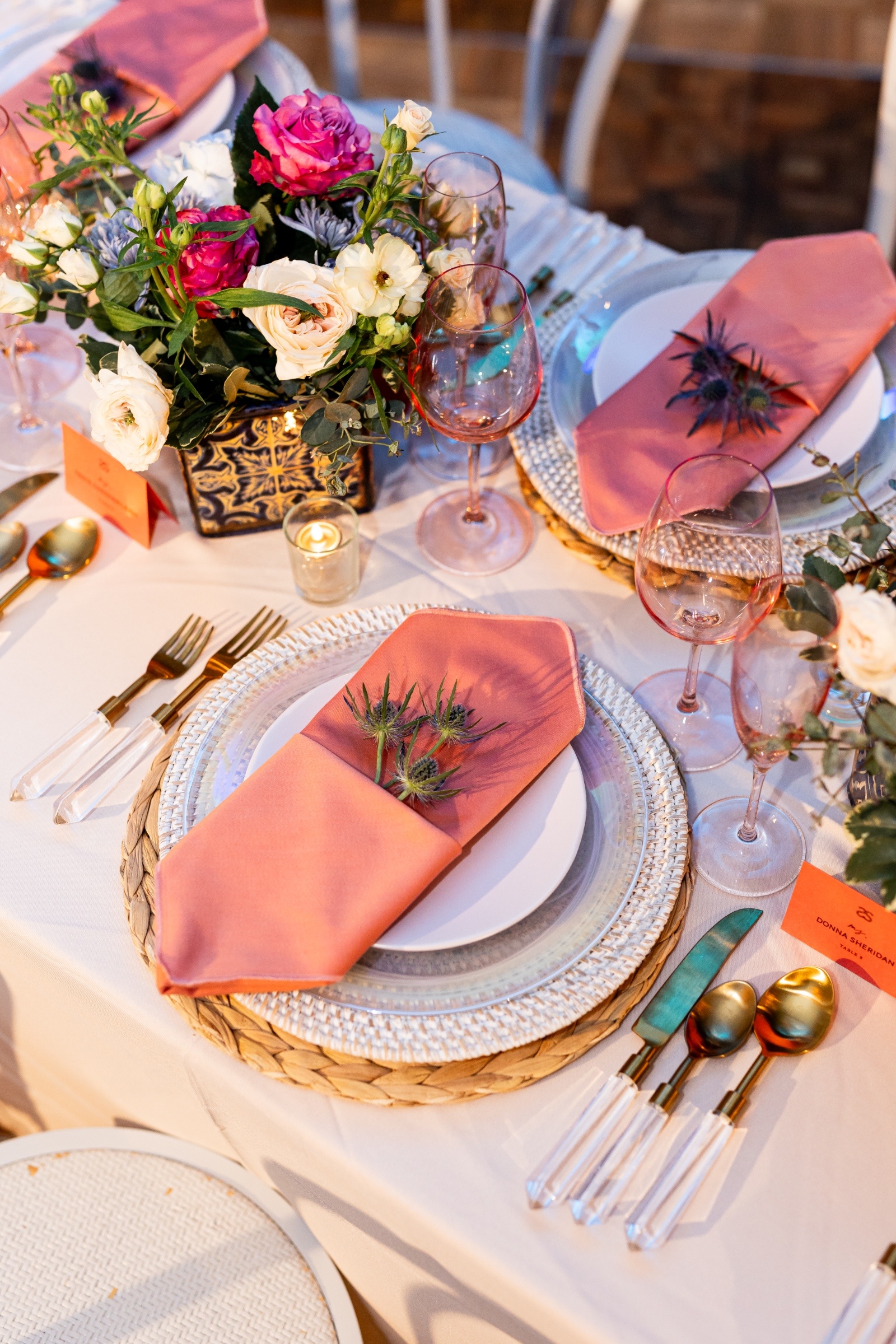 Elegant table setting with peach napkins adorned with sprigs on white plates, crystal cutlery, and pink wine glasses. A vibrant floral centerpiece adds a fresh touch.